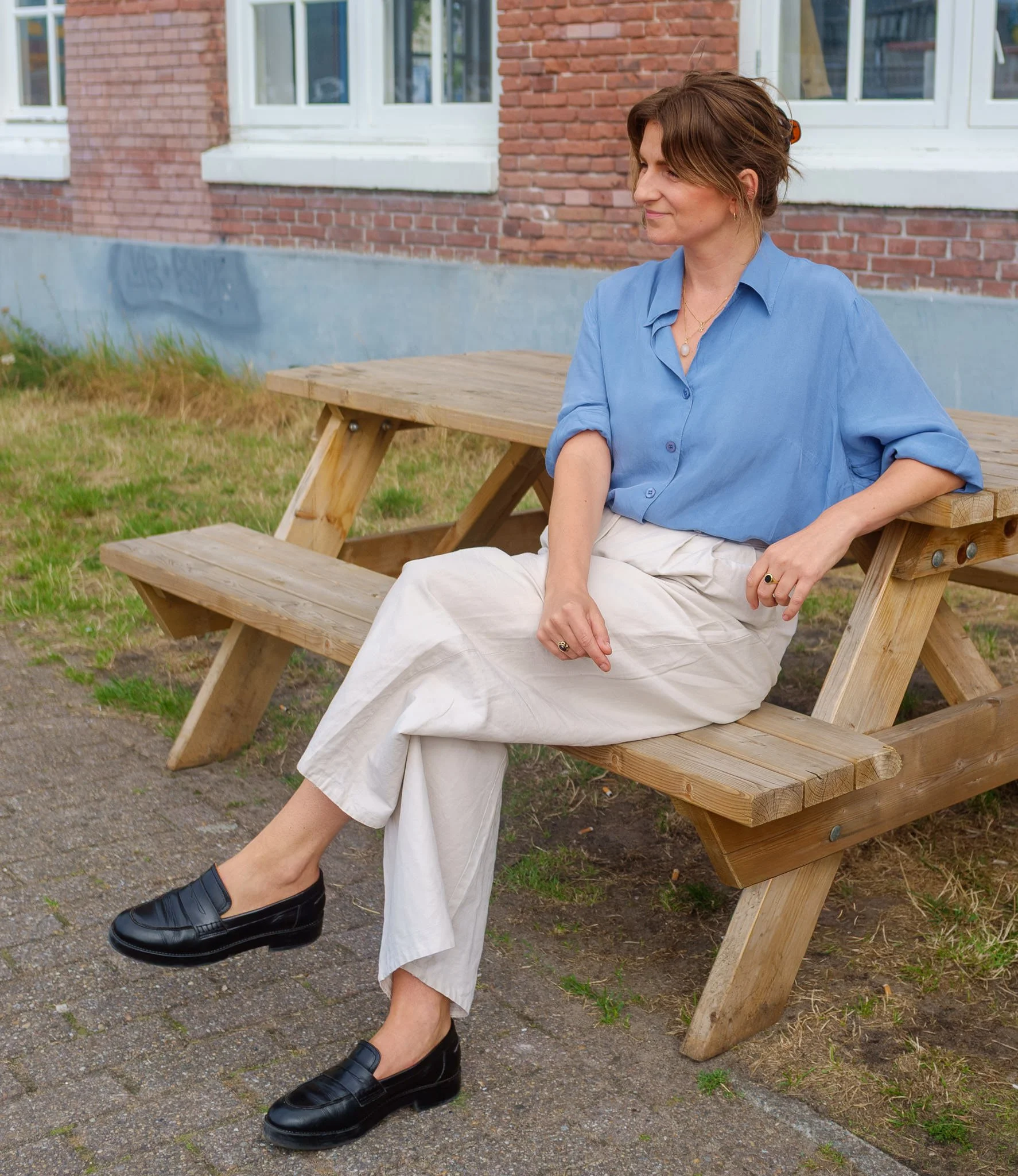 een vrouw met schouderwolk haar zit op een houten bank buiten voor een bakstenen gebouw, draagt een lichtblauwe blouse, witte broek, zwarte schoenen, en draagt sieraden.