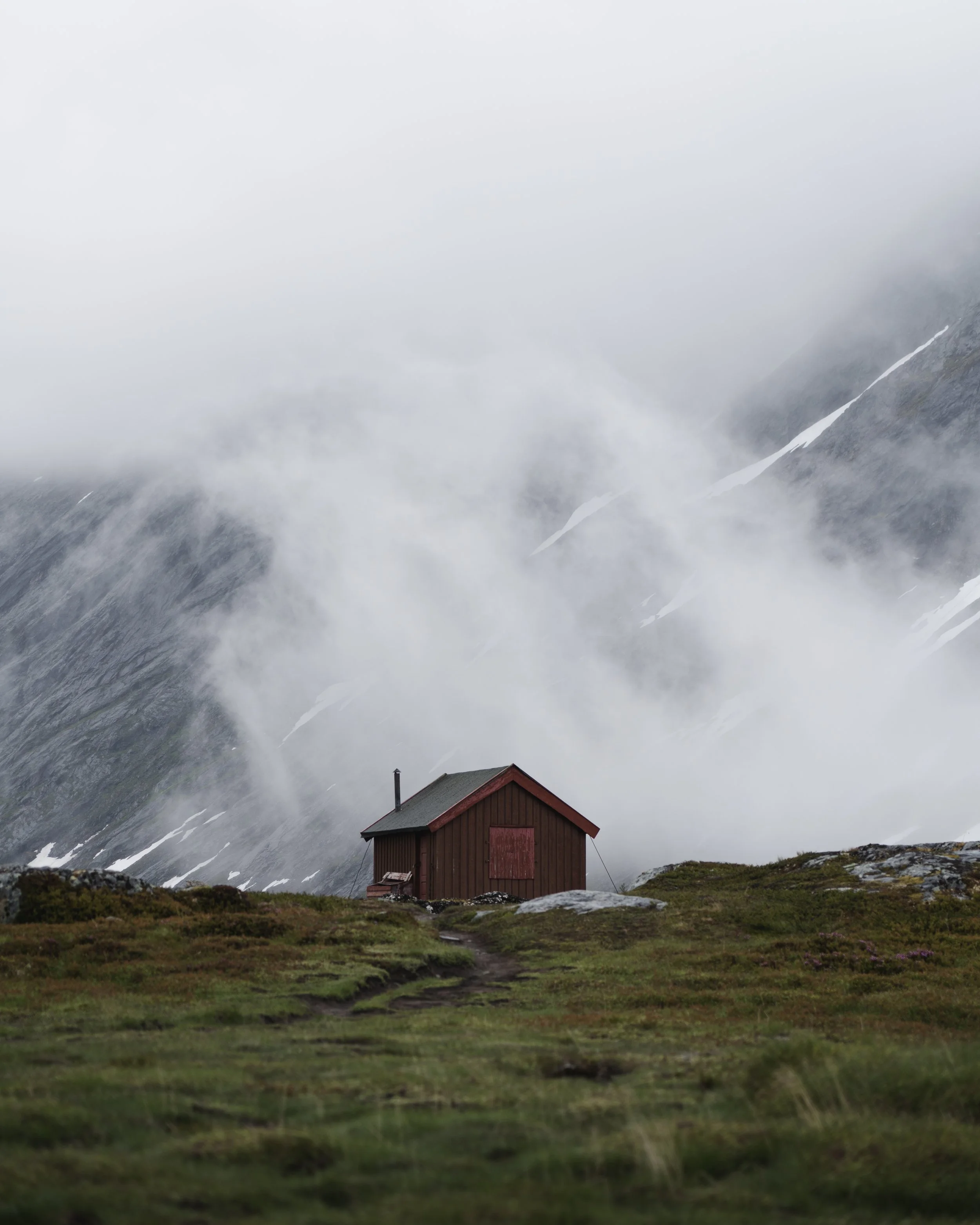 Ein kleines rotes Haus auf einer grünen Wiese vor einer nebelverhangenen Bergkulisse mit Schnee an den Hängen.