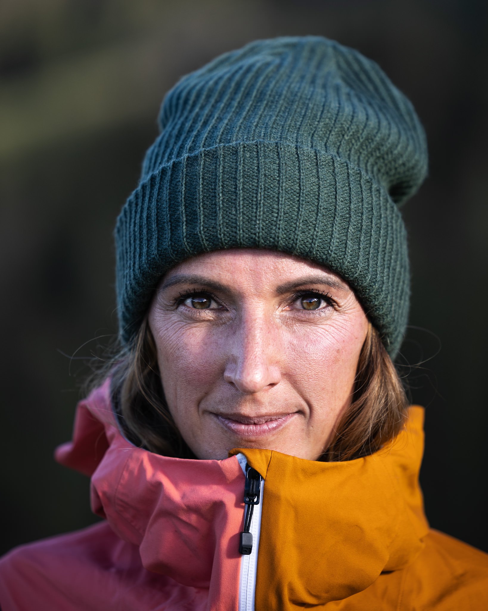 Nahaufnahme eines Mannes mit braunen Haaren und grünen Augen, trägt einen grauen Strickmütze und eine Outdoor-Jacke in Orange und Rosa, blickt zur Kamera, vor unscharfem dunklen Hintergrund.