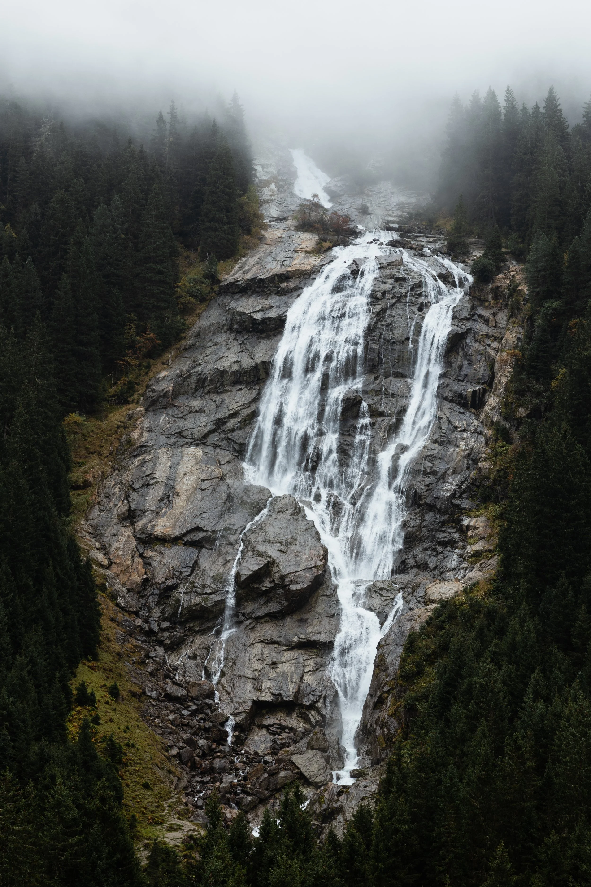 Ein Wasserfall in einer bewaldeten Bergregion, umgeben von Tannenbäumen, an einem nebligen Tag.