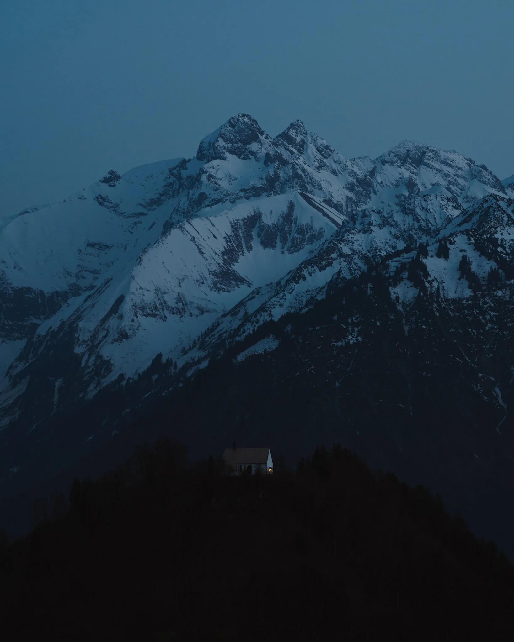 Schneebedeckte Berge bei Nacht mit einem kleinen Haus mit Licht im Vordergrund.