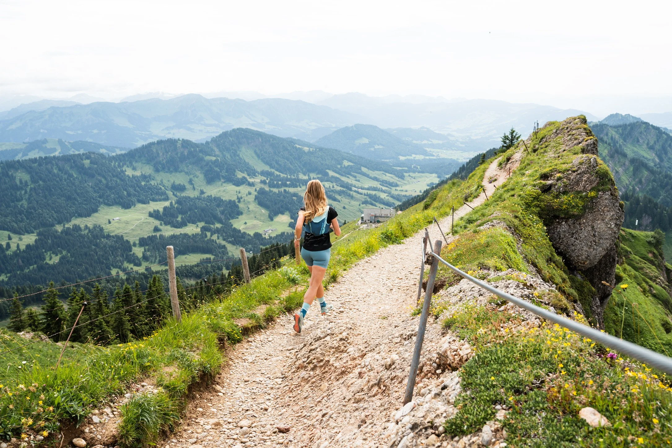 Eine Frau beim Wandern auf einem Bergpfad in den Alpen, umgeben von grünen Hügeln und Bergen, mit einem blauen Rucksack und sportlicher Kleidung.