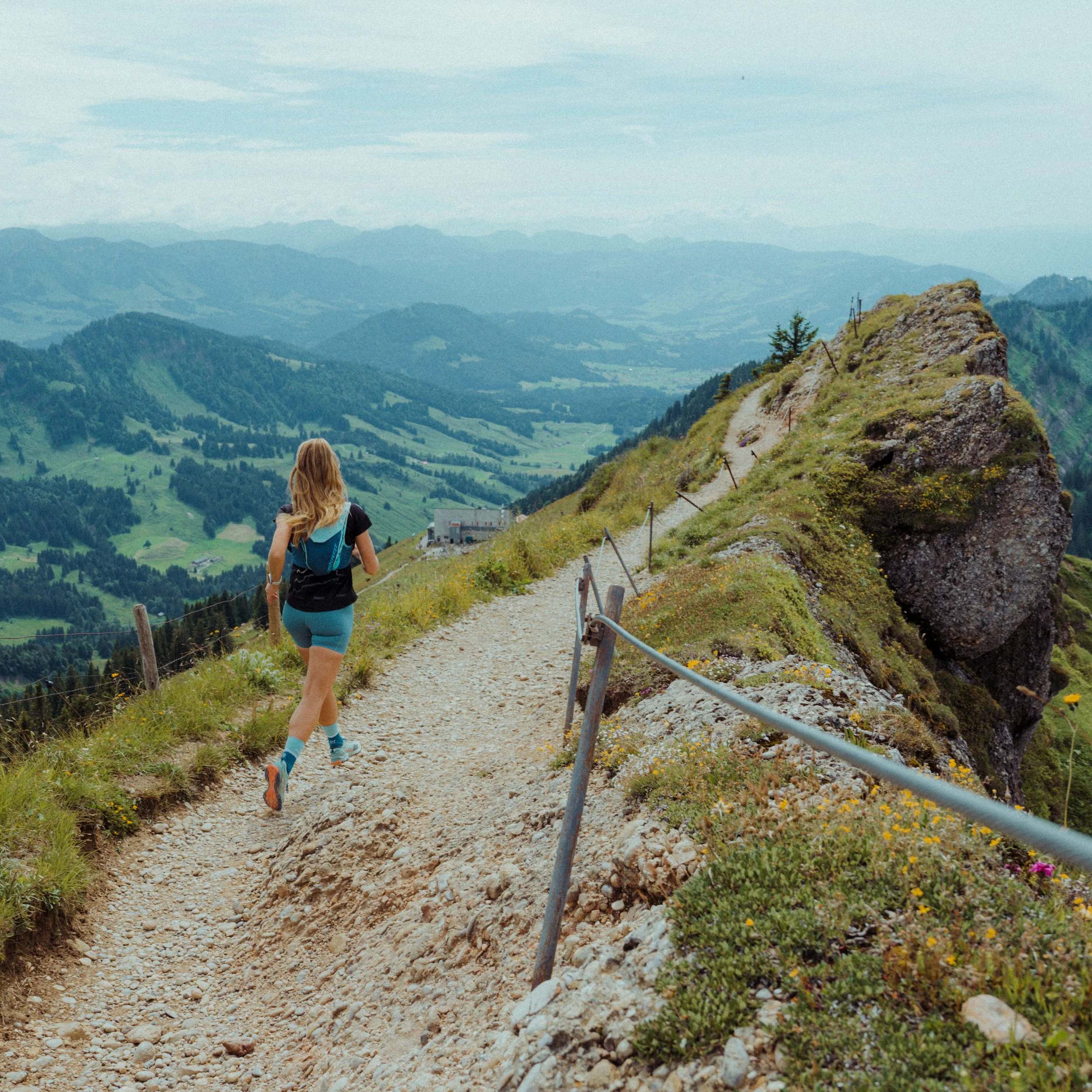 Eine Frau läuft auf einem schmalen Wanderweg entlang eines Hügels mit grüner Vegetation und bunten Blumen, in einer bergigen Landschaft mit mehreren Hügeln und Bergen im Hintergrund.