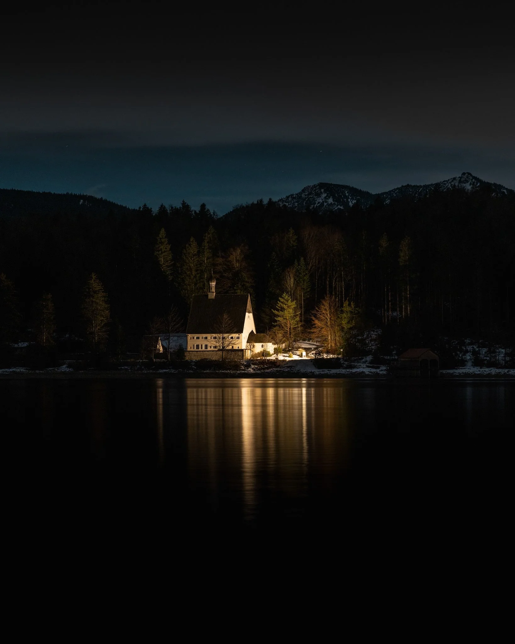 Abendstimmung an einem See mit Haus und Bergen im Hintergrund, reflektiert im Wasser.