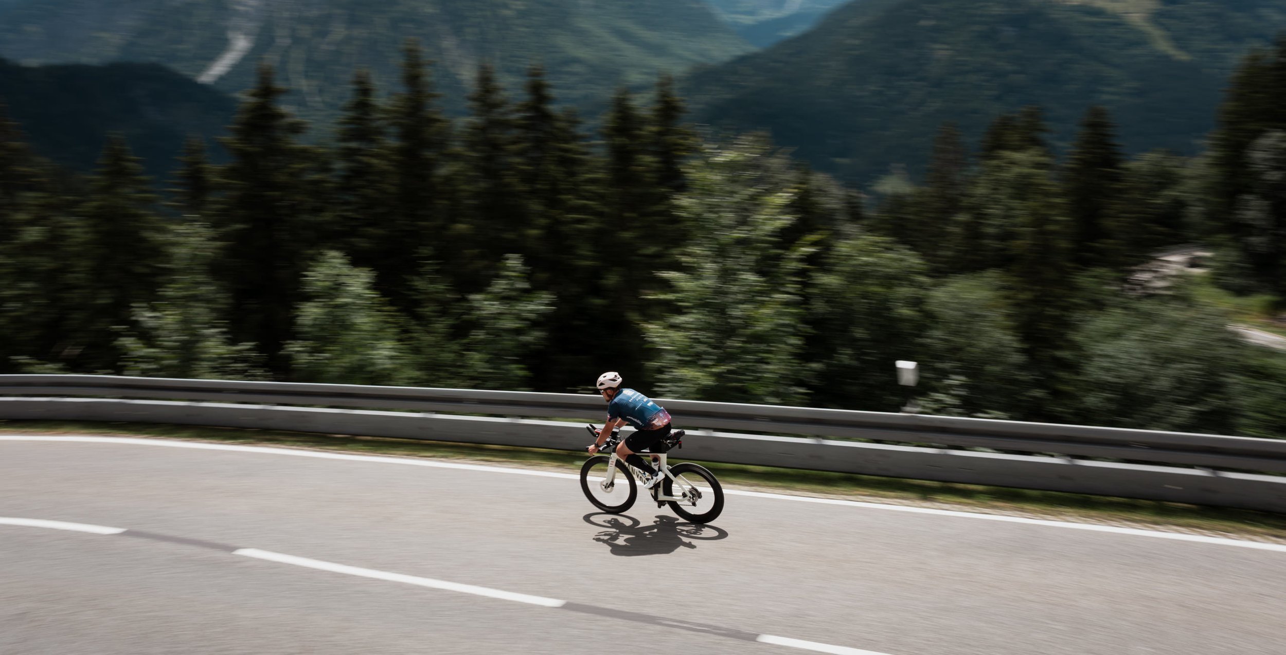 Ein Radfahrer fährt auf einer kurvenreichen Straße an einem bewaldeten Gebirge vorbei.