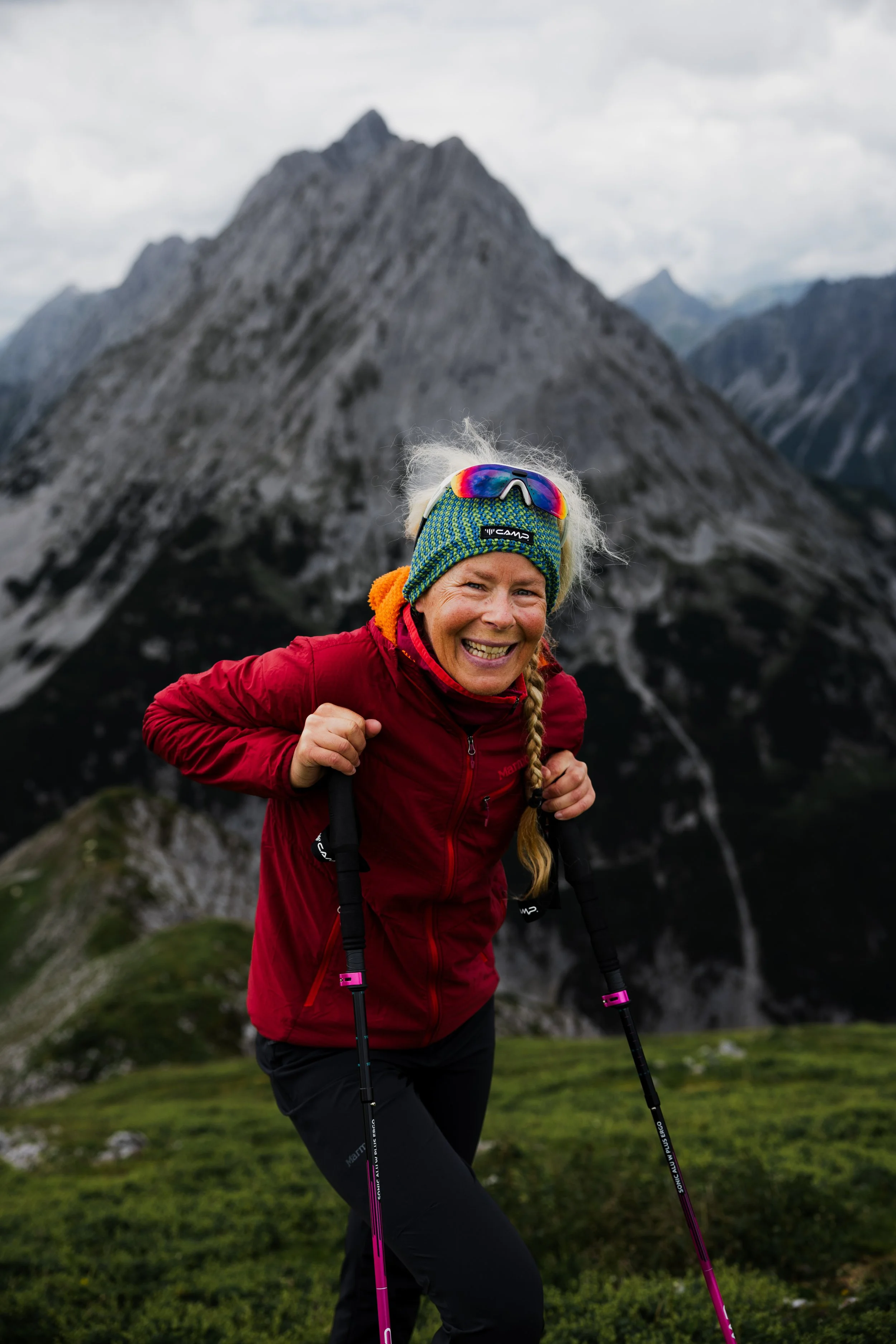 Frau beim Bergwandern im Gebirge, mit grünem Beanie, rote Jacke und Skistöcken, vor einer Bergkette unter Wolken am Himmel.