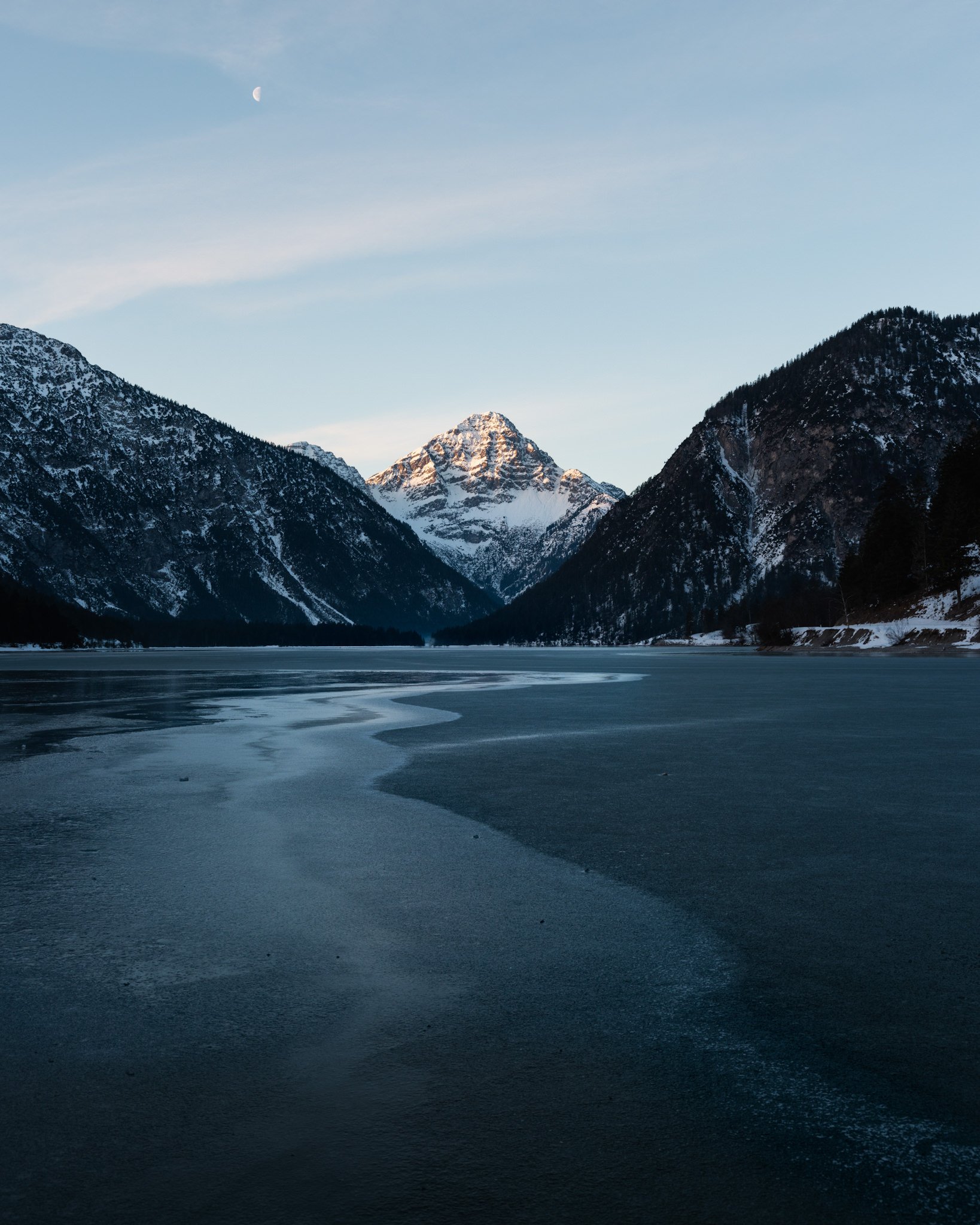 Berglandschaft mit schneebedeckten Gipfeln, einem gefrorenen See im Vordergrund, und einem Halbmond am Himmel.