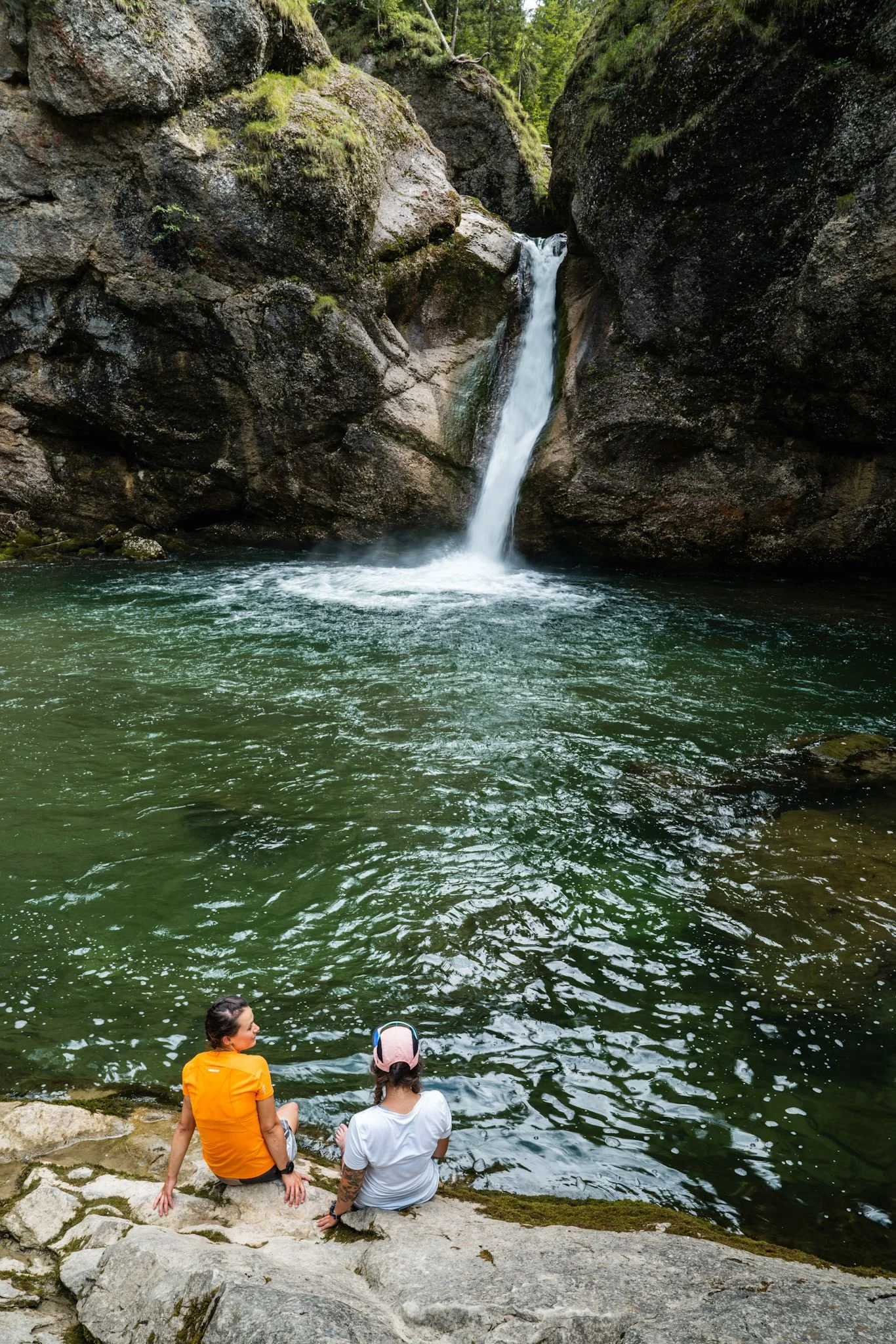 Drei Personen sitzen am Ufer eines Flusses, im Hintergrund ein Wasserfall in einer bewaldeten Schlucht.