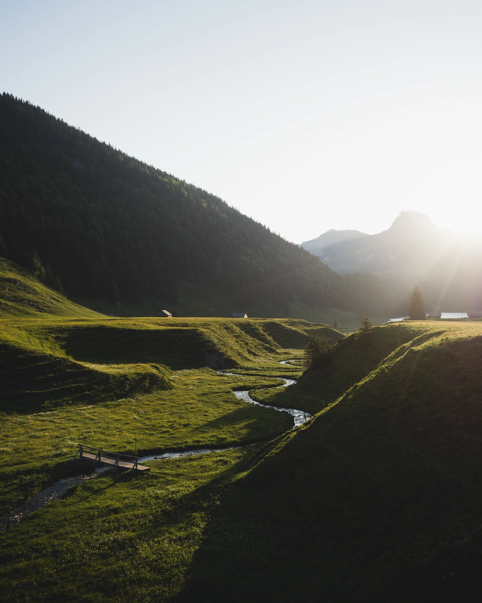 Eine ländliche Landschaft mit grünem Tal, einem kleinen Fluss und Bergen im Hintergrund, beleuchtet von Sonnenlicht.