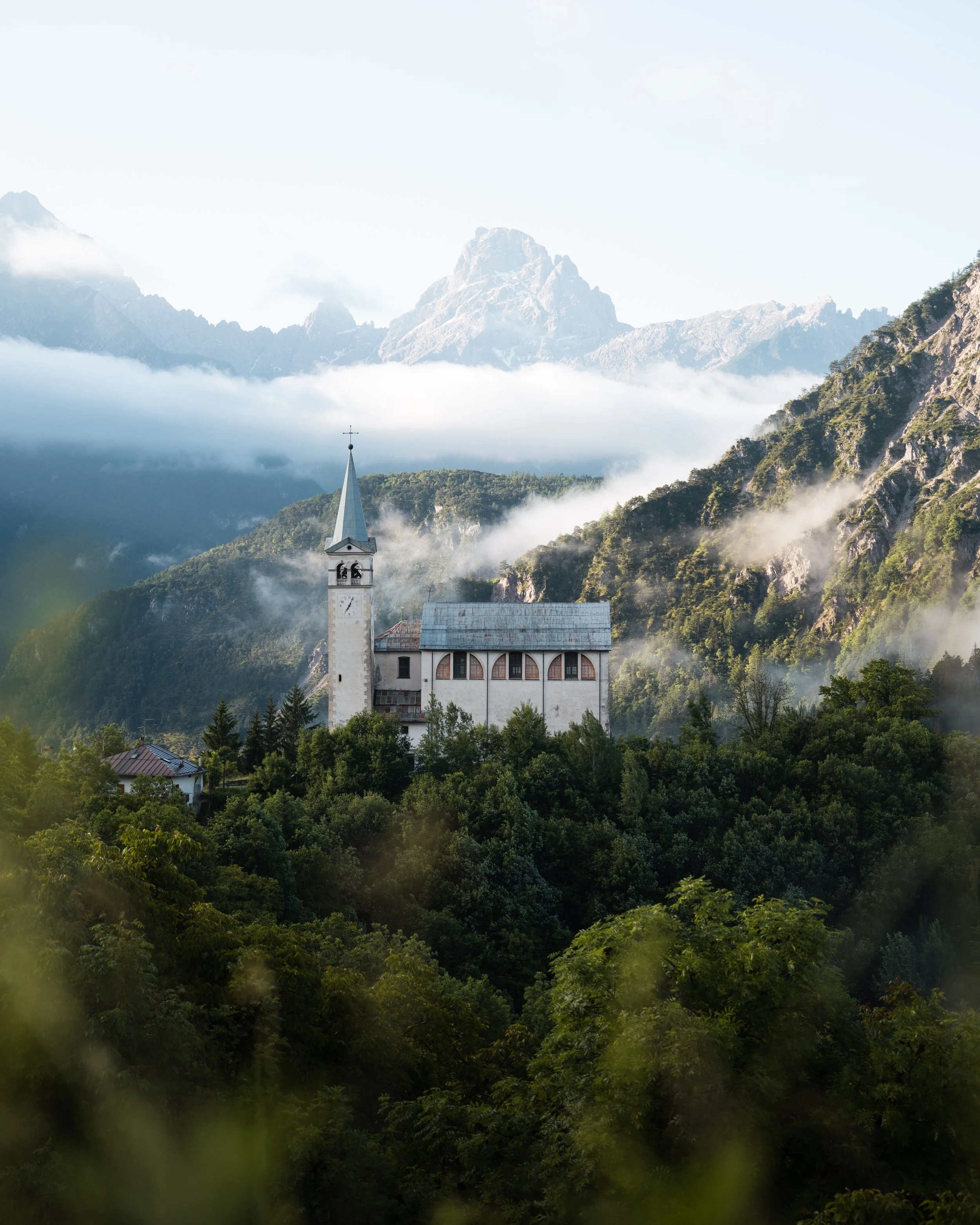 Ein malerischer Bergsee mit hohen, schneebedeckten Gipfeln im Hintergrund, umgeben von üppigem Grün und einer kleinen weißen Kirche im Vordergrund.