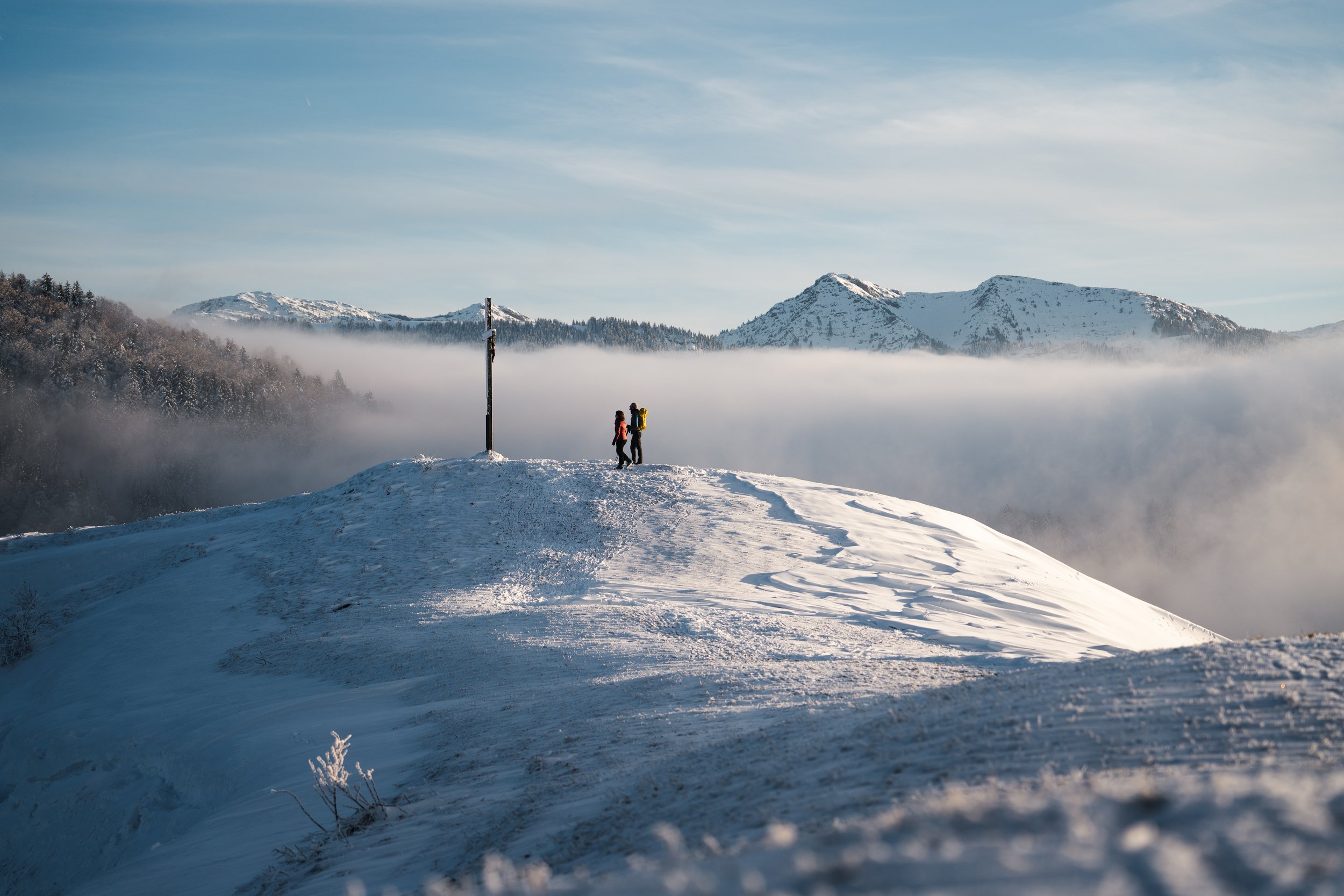 Zwei Wanderer mit Rucksäcken auf schneebedeineter Bergkuppe, im Hintergrund verschneite Berge und Nebel