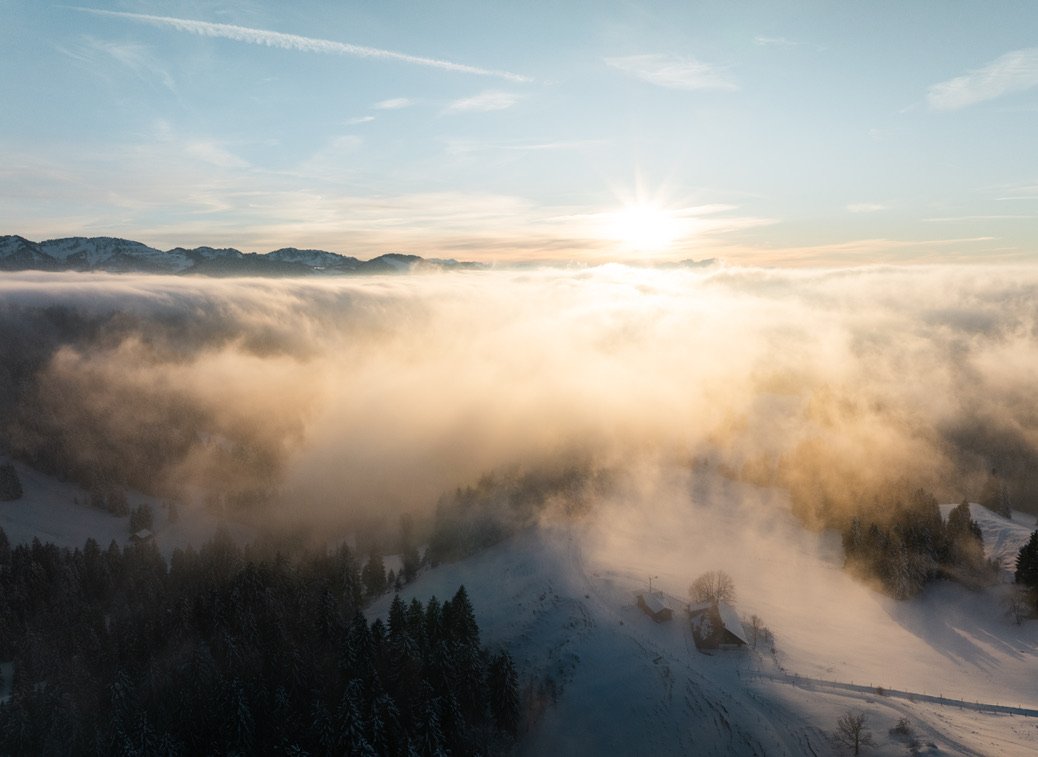 Blick auf eine verschneite Berglandschaft mit Nebel und Wolken, Sonnenaufgang am Horizont.