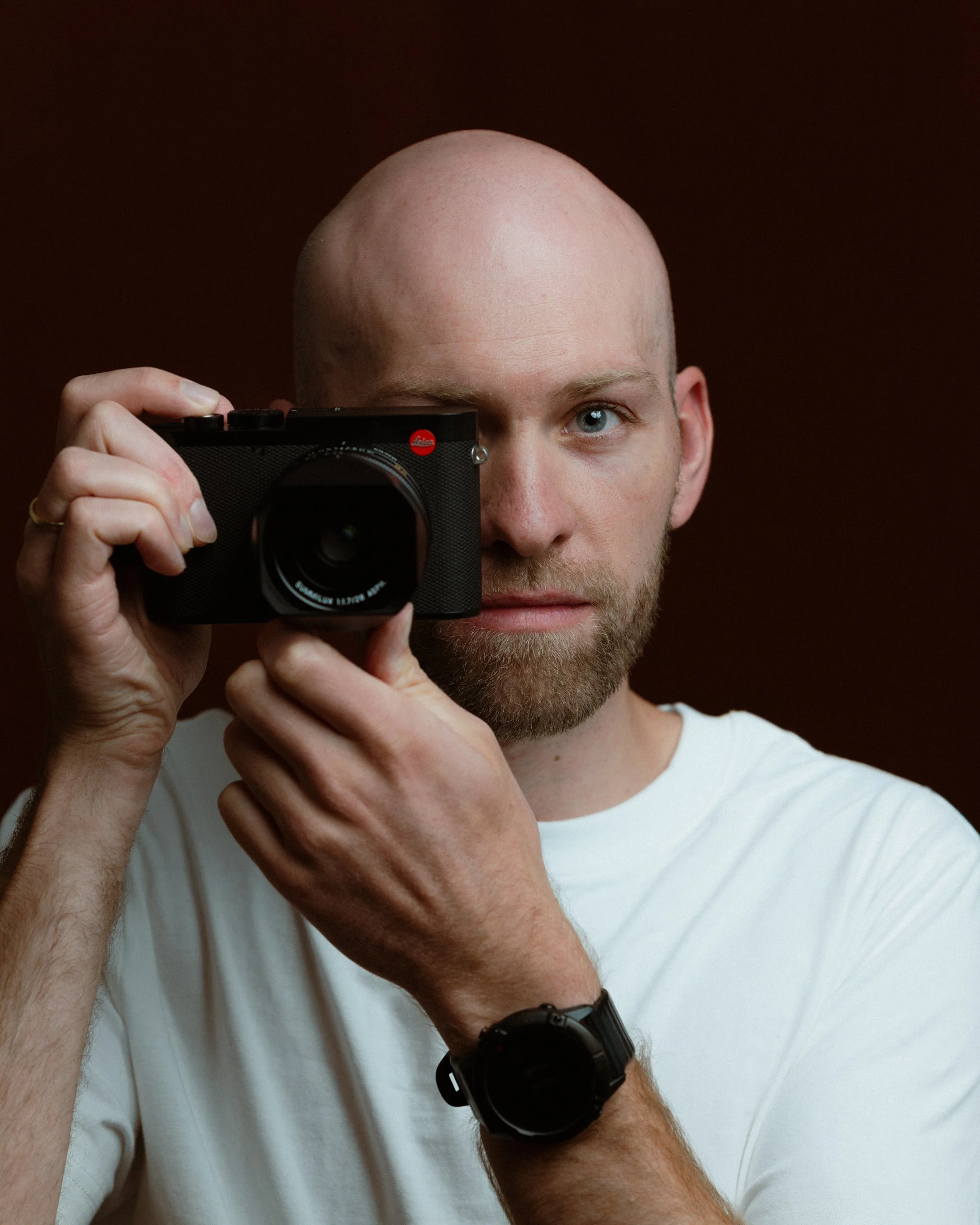 Der Fotograf Simon Brausten mit blonden Haaren und blauer Augen hält eine Leica Kamera vor sein Gesicht. Er trägt eine weiße T-Shirt und eine schwarze Uhr.