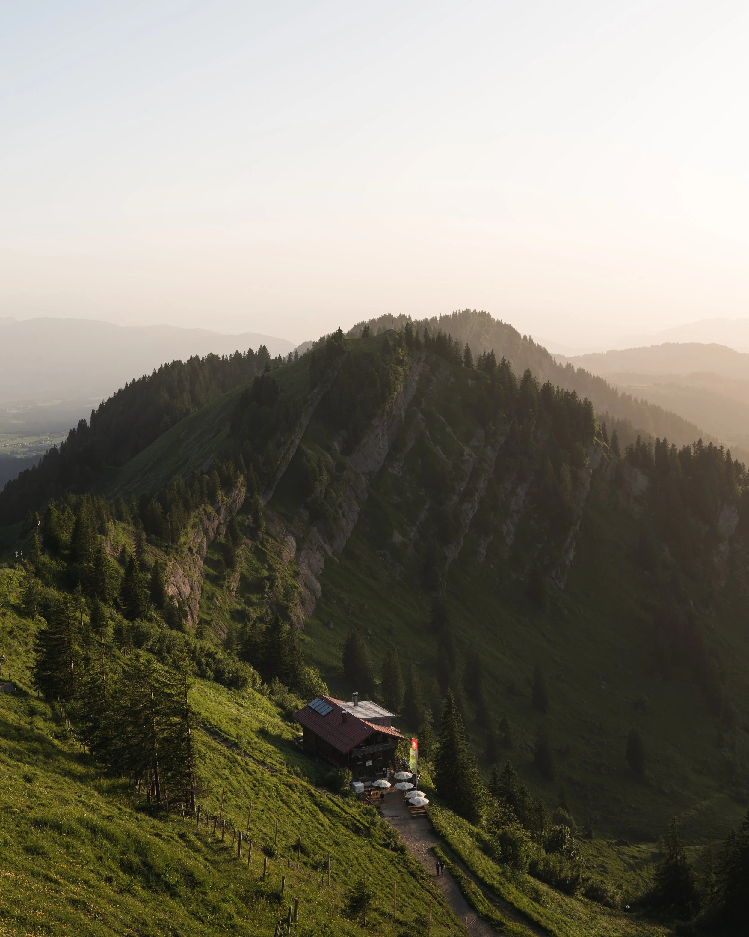 Ein kleines Haus mit Terrasse und Sonnenschirmen, umgeben von grünen Hügeln und Bergen bei Sonnenuntergang.