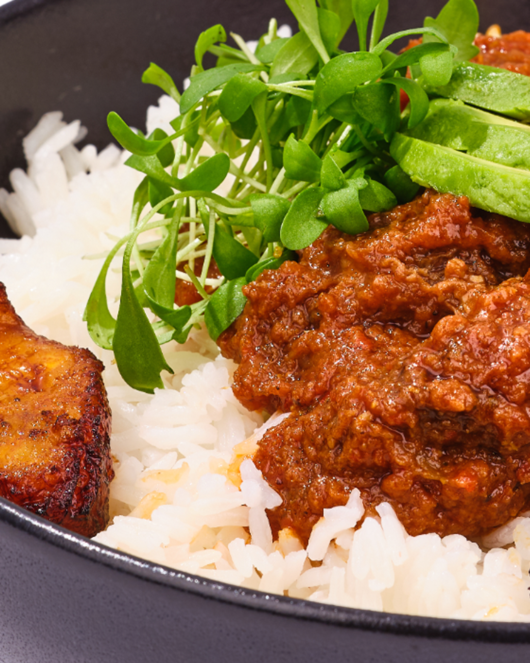Close-up of a sophisticated black bowl featuring fluffy white rice, a tender meat richly coated in a vibrant tomato sauce, fresh crisp greens, and an elegant slice of fresh avocado.