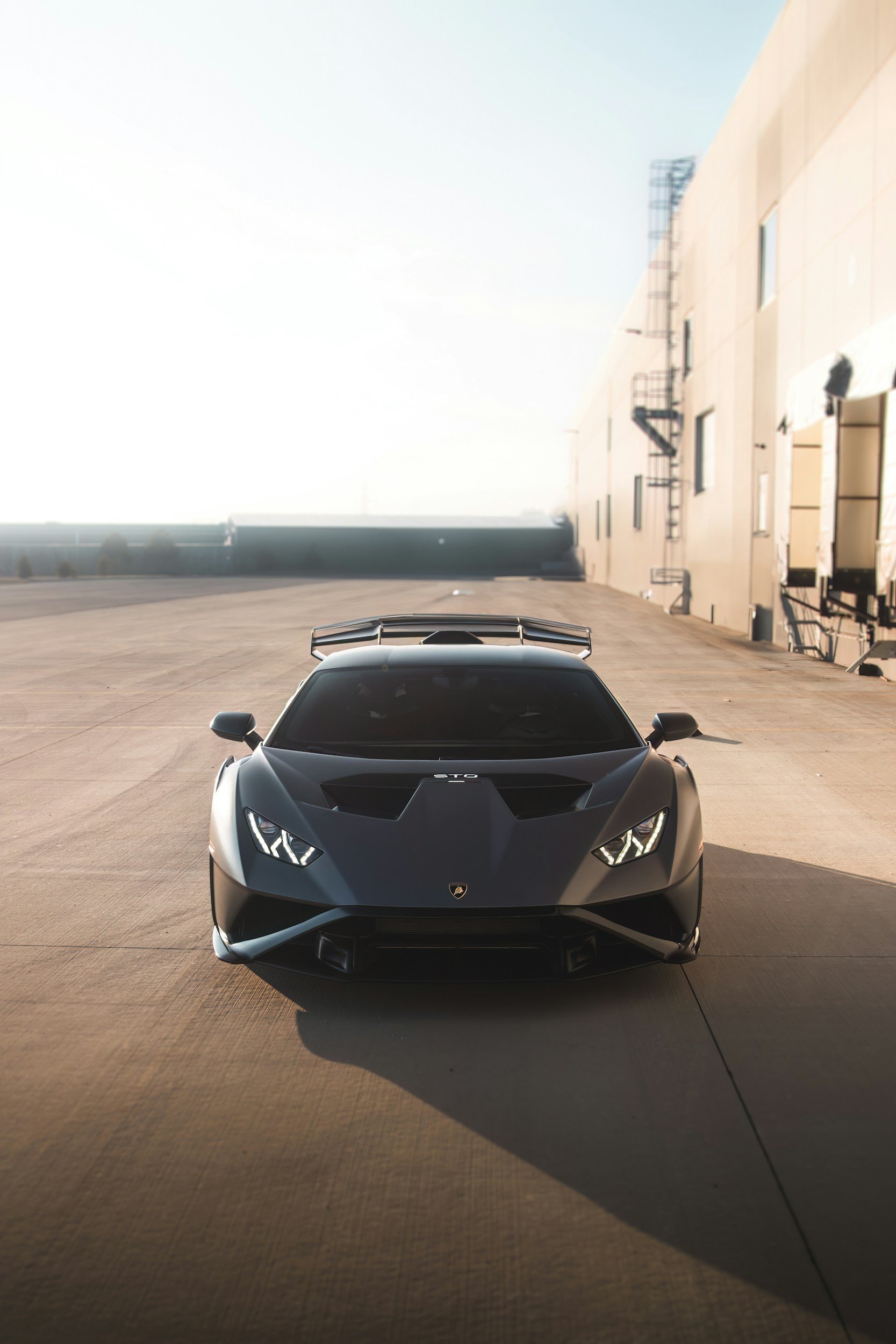 Front view of a matte black Lamborghini sports car parked outdoors, with a beige building wall with windows and metal fire escapes on the right and a bright sky in the background.