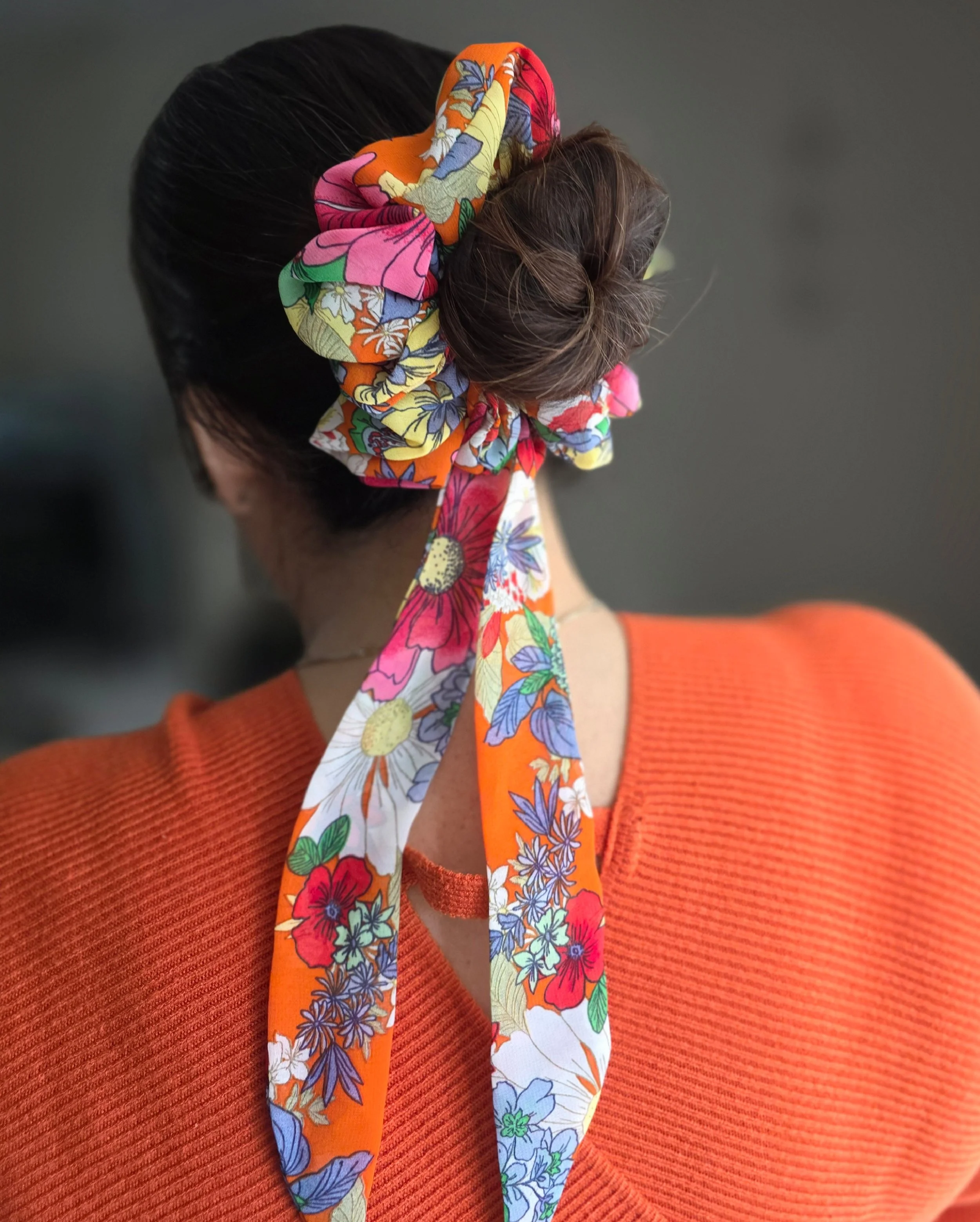 A person with dark hair styled in a bun, wearing a bright orange sweater, has a colorful floral fabric scarf tied around their head in a bow, viewed from behind.