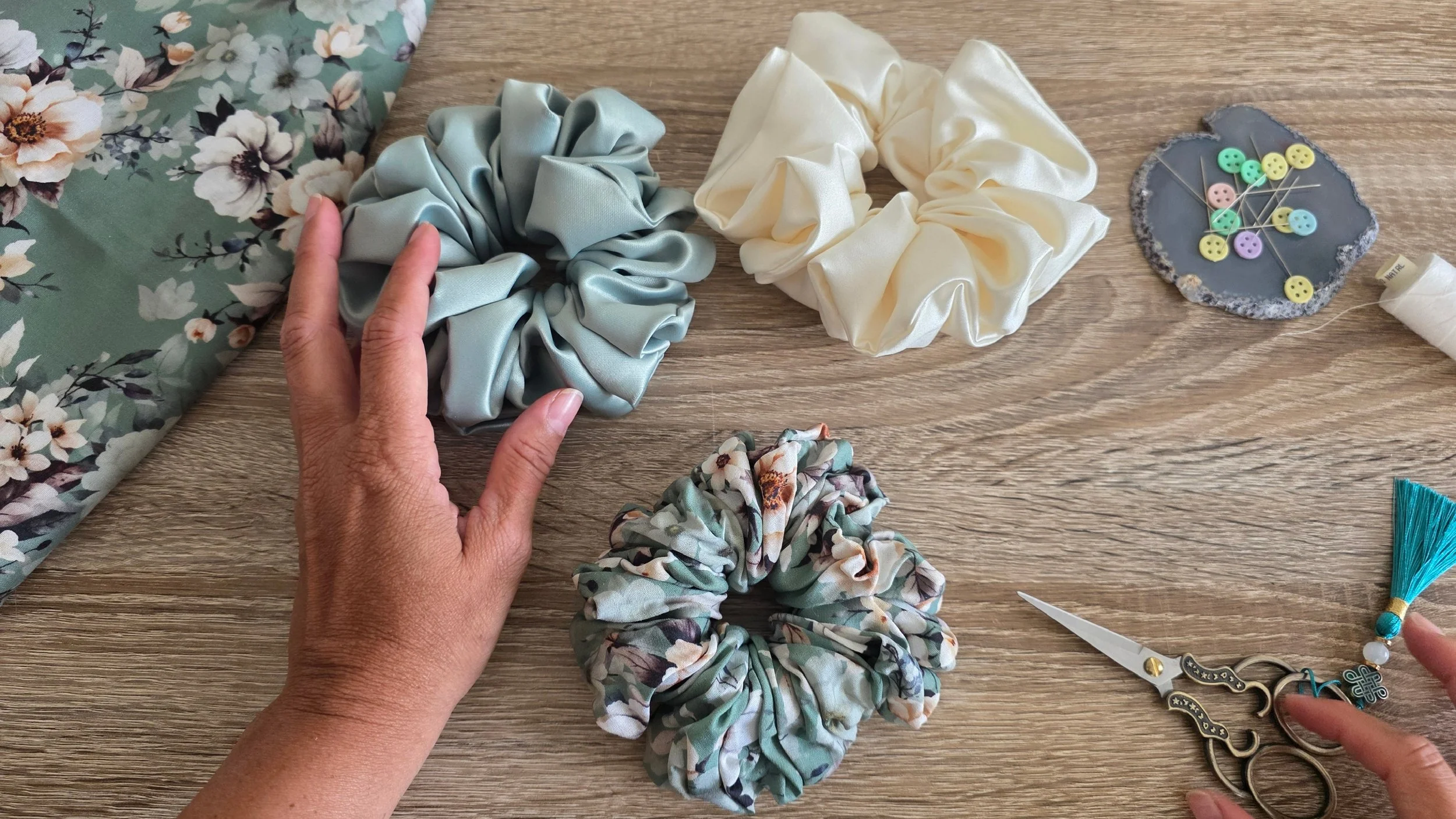 A pair of person's hands arranging fabric scrunchies on a wooden surface with sewing tools and accessories nearby.