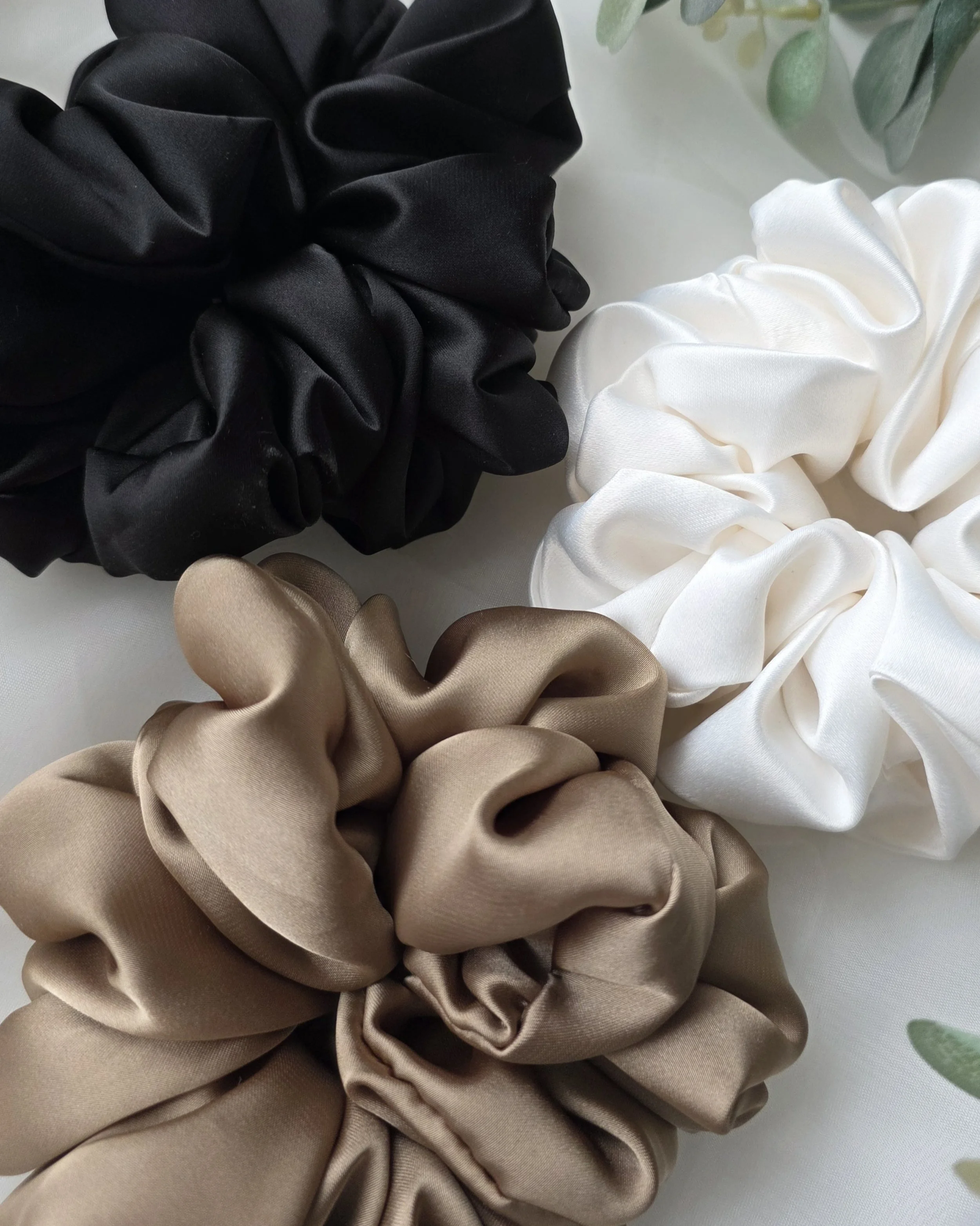 Close-up of fabric scrunchies in black, beige, white, and a hint of tan colors, arranged on a light-colored surface with a partial view of a plant in a pot in the top right corner.