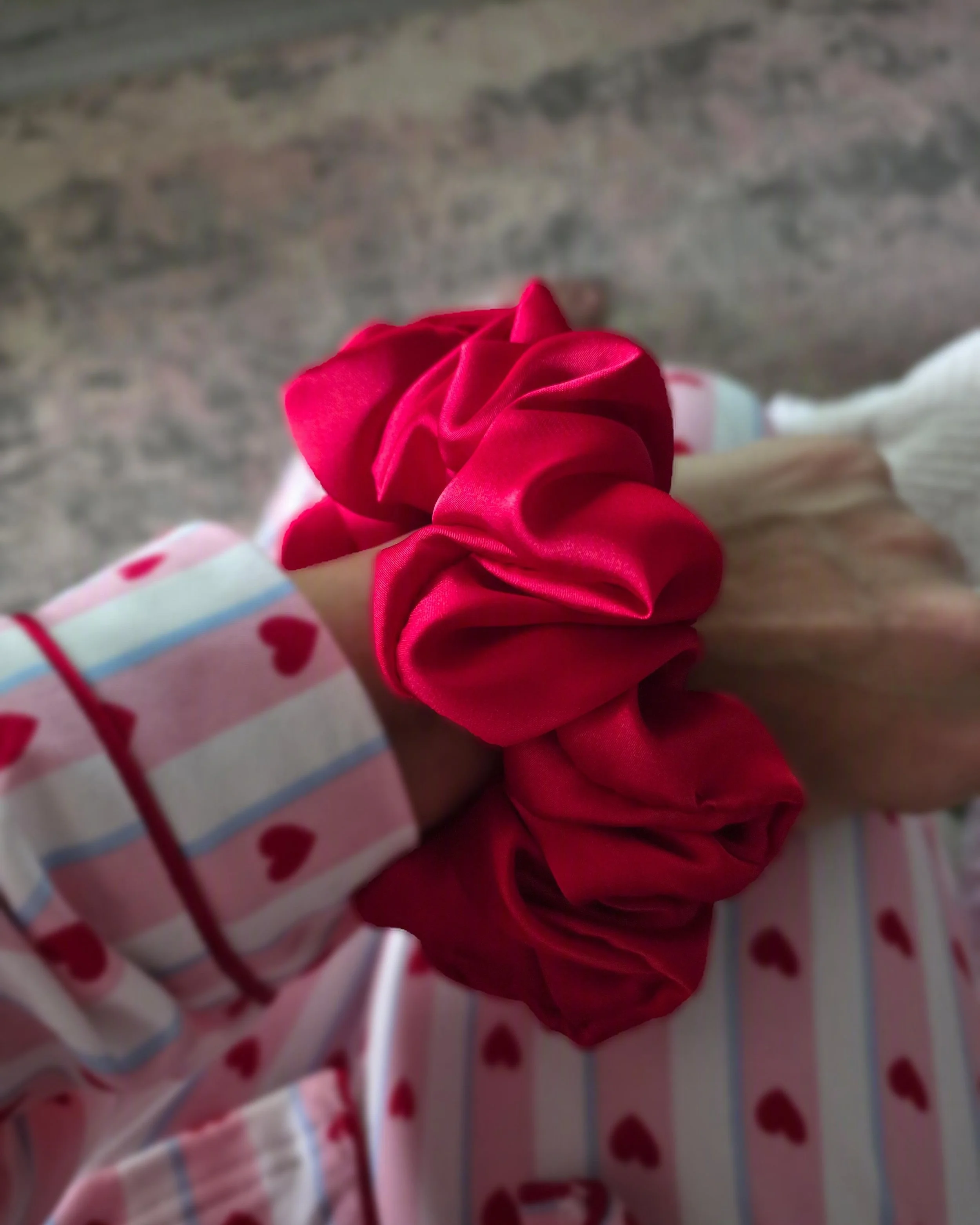 A person holding a red satin scrunchie. The person is wearing pajamas with red hearts and light blue stripes on a white background. The background appears to be a concrete or stone surface.