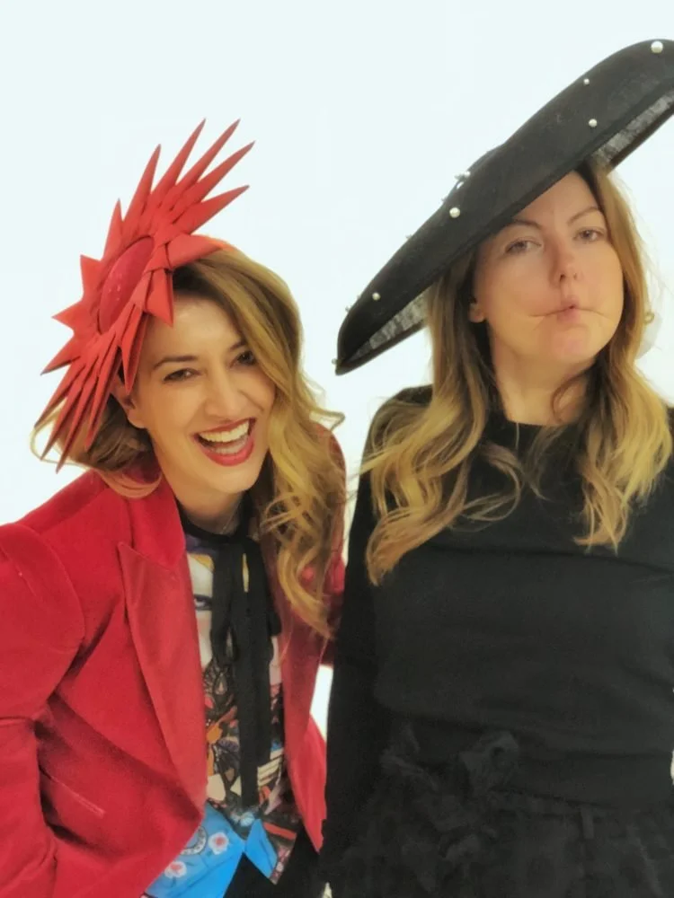 Two women wearing festive hats, one in a red jacket with a red feathered hat and the other in black with a wide-brimmed black hat, smiling and posing for a photo.