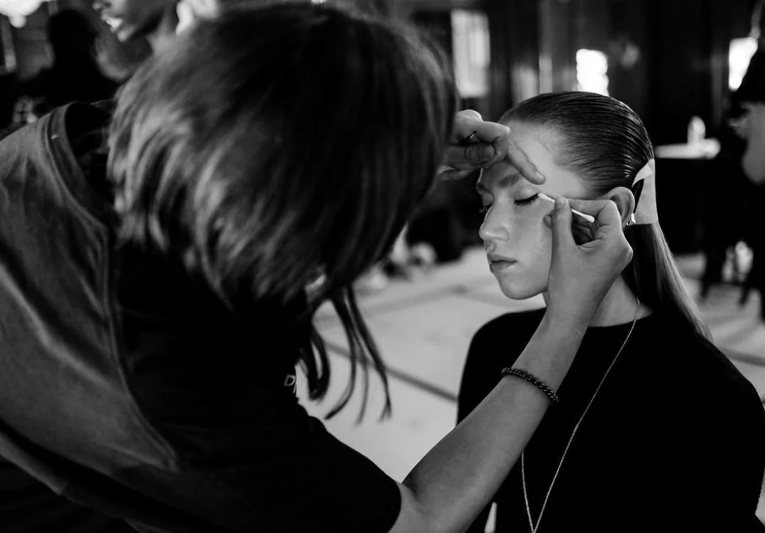 A makeup artist applying eye makeup to a young woman with slicked-back hair in a backstage setting.