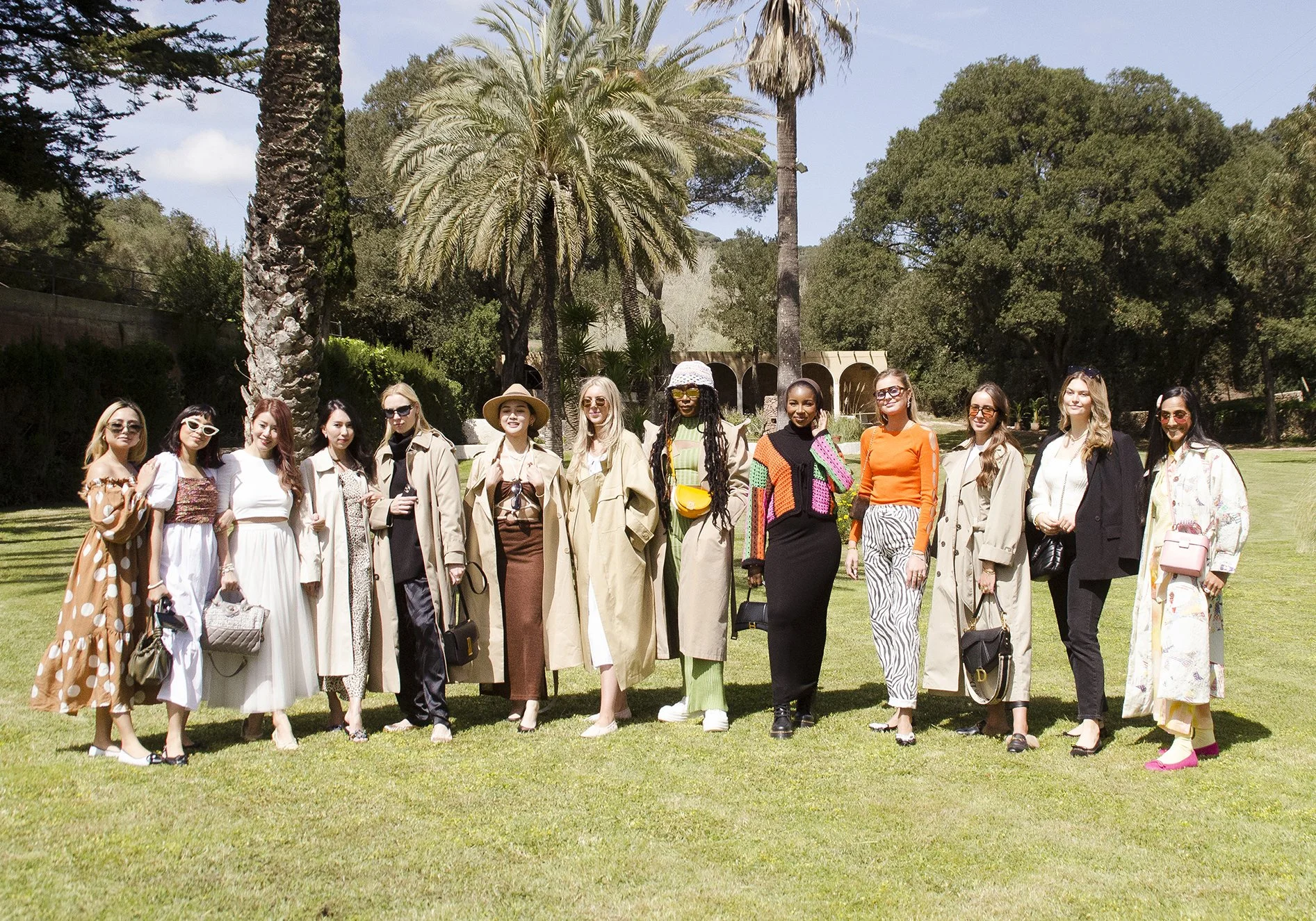 A group of women dressed in fashionable and colorful outfits standing outdoors on a lush green lawn with palm trees and other trees in the background on a sunny day.