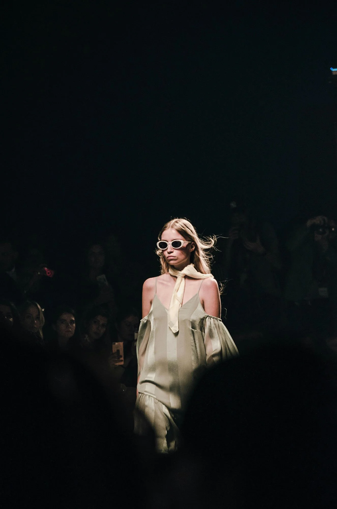 Model walking down runway in beige outfit with cold shoulder design, sunglasses, and scarf, during fashion show with audience.