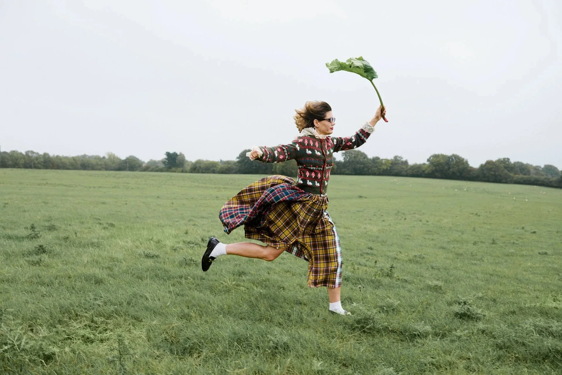 A woman running outdoors in a grassy field while holding a large leaf and wearing a colorful sweater and plaid skirt.