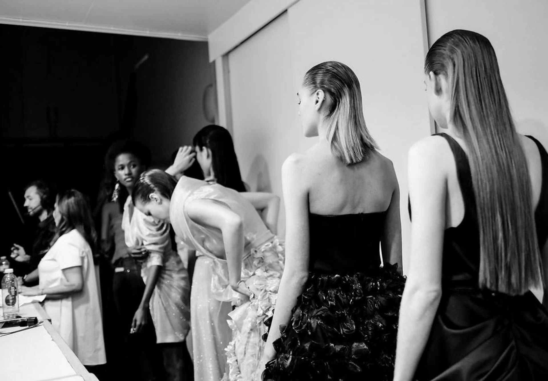 A black-and-white photo of women getting ready backstage, some are dressed in elegant outfits, with others in casual and formal attire, preparing for a performance or event.