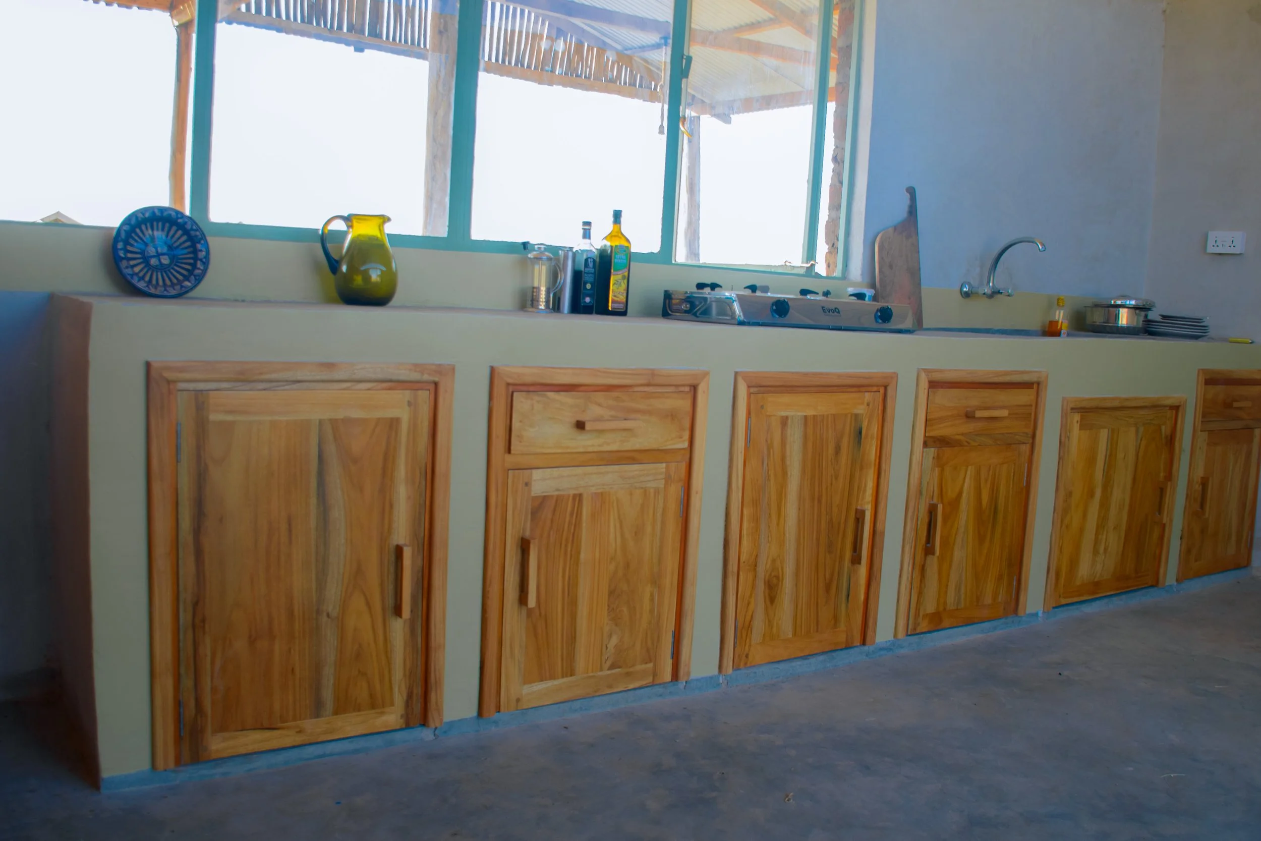 Kitchen with wooden cabinets, a countertop, and various bottles and kitchen items, located near a large window with a view of a covered outdoor area, at Njiani River Camp Moshi