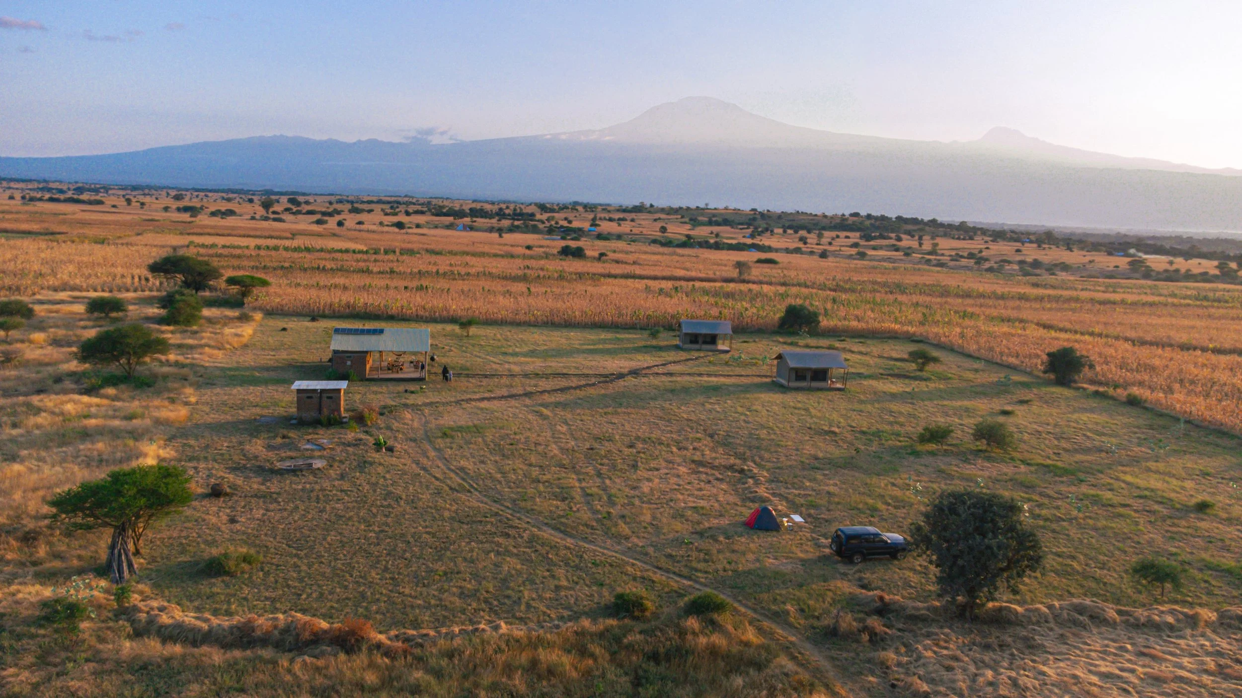 A wide landscape view of an open field near Moshi with several small buildings, a tent, and a vehicle, with Kilimanjaro in the background.