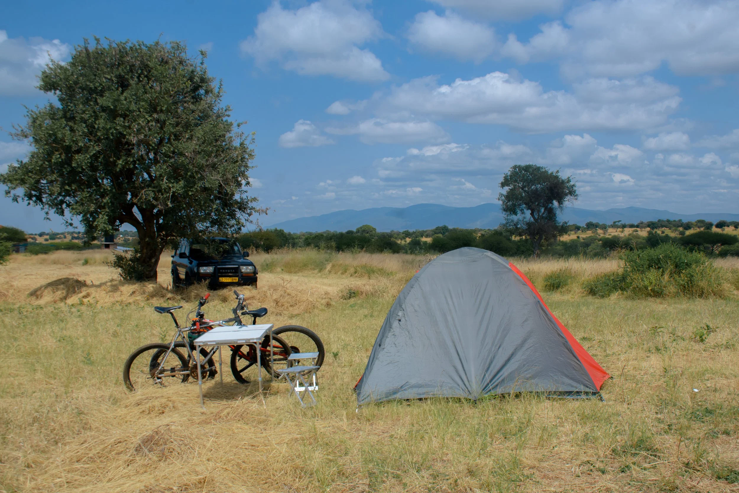 Camping scene with a tent, bicycles, and a car in an open field under a partly cloudy sky, with Kilimanjaro in the background.