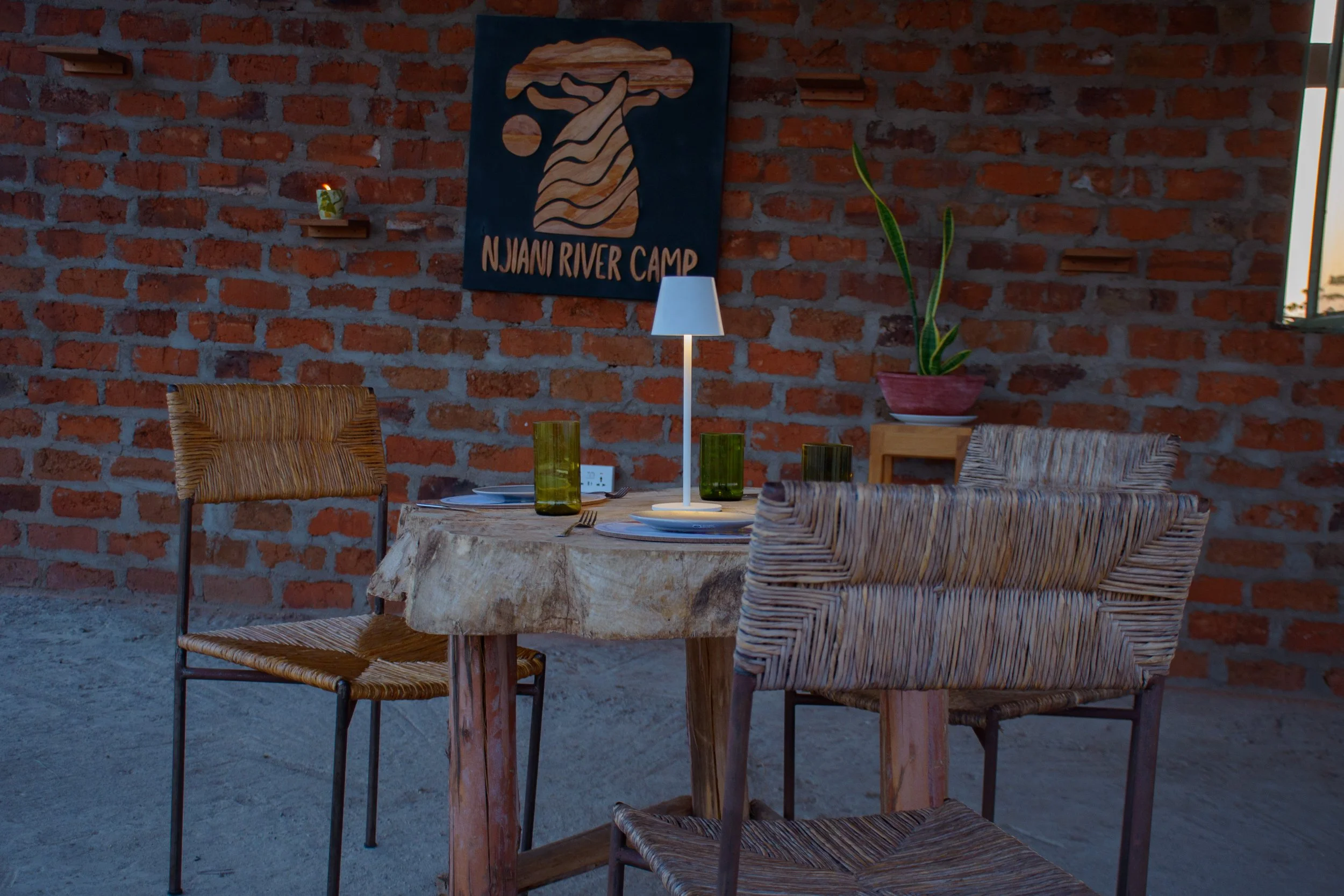 A rustic dining area with a wooden table set with plates, green glasses, and a small lamp. Wicker chairs surround the table. The background features a brick wall with a blue sign that reads NJIANI RIVER CAMP.