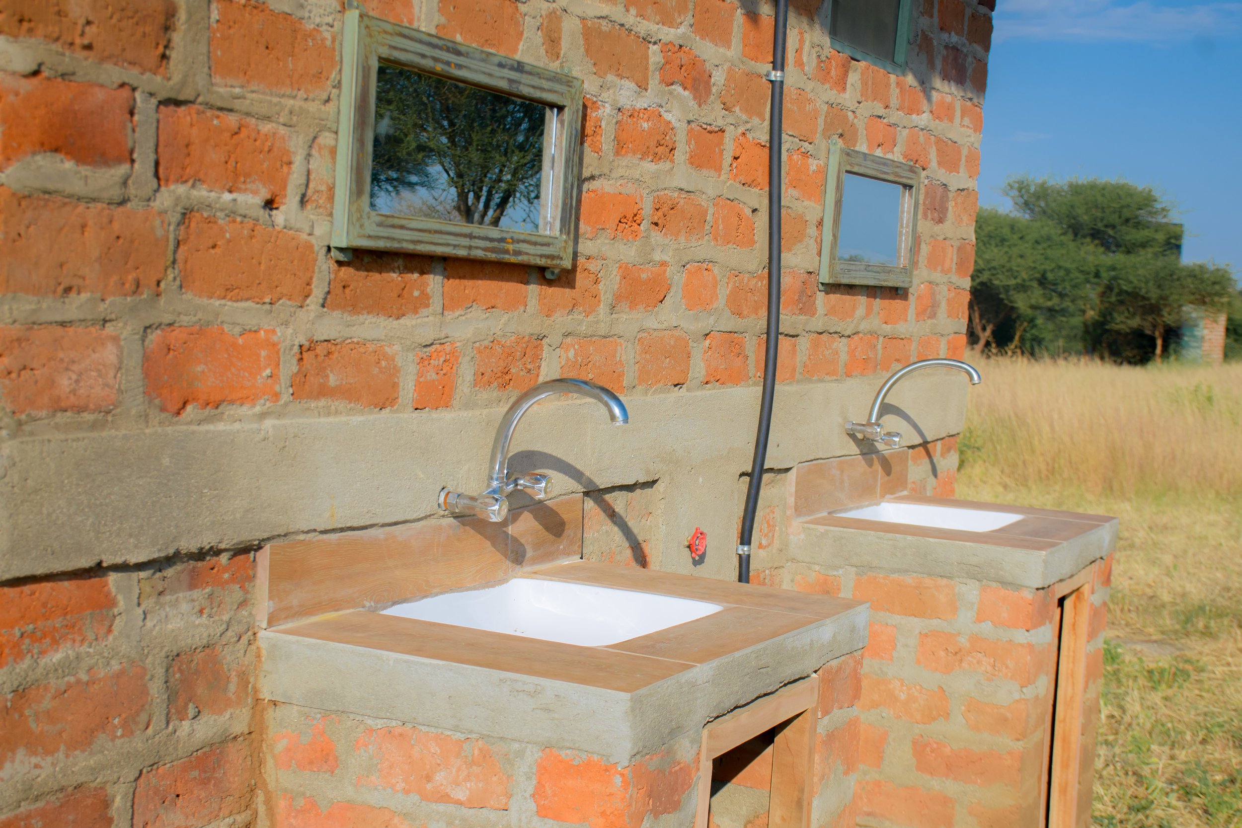 Outdoor brick wall with two small sinks and faucets, mirrors on the wall, and a grassy field with trees in the background.