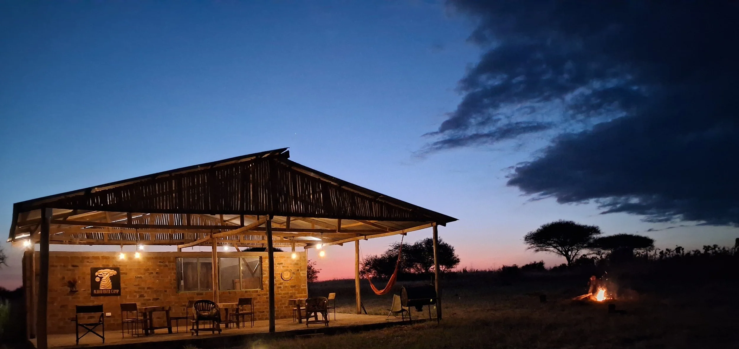 A rustic outdoor structure, at Njani River Camp, with a thatched roof, string lights, and chairs, set against a sunset sky with a few clouds and trees in the background. There's a campfire on the right side.
