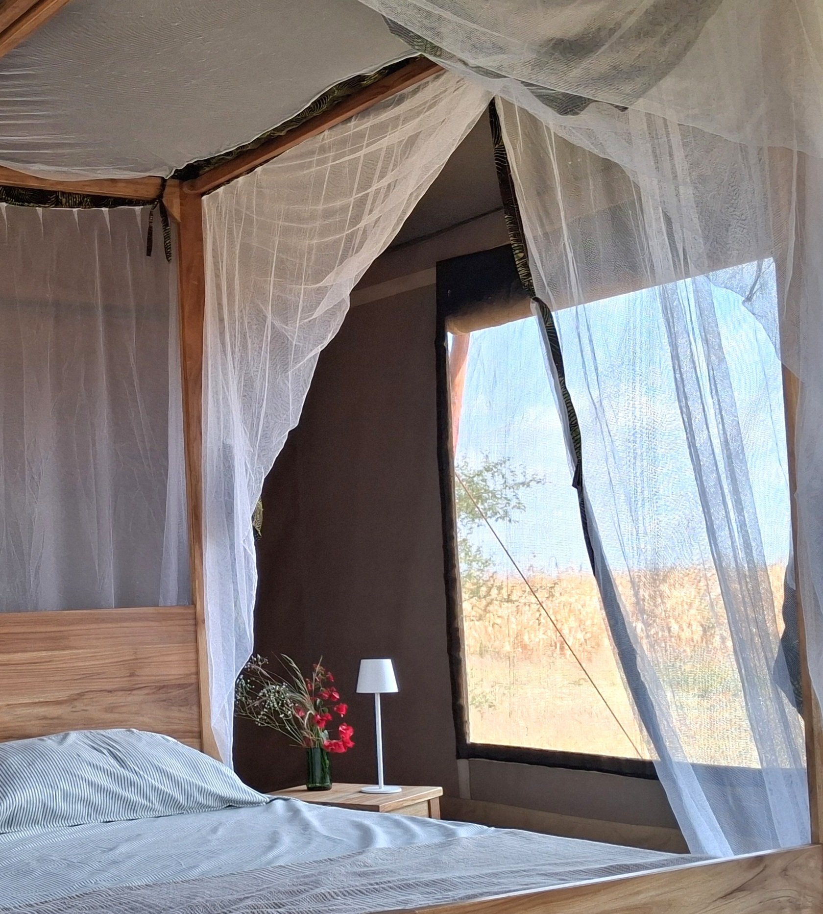 Interior of a tent with a wooden bed frame and white canopy drapes, two beds with white sheets, dark curtains on the sides, and small nightstands with lamps at Njiani Camp, Moshi.