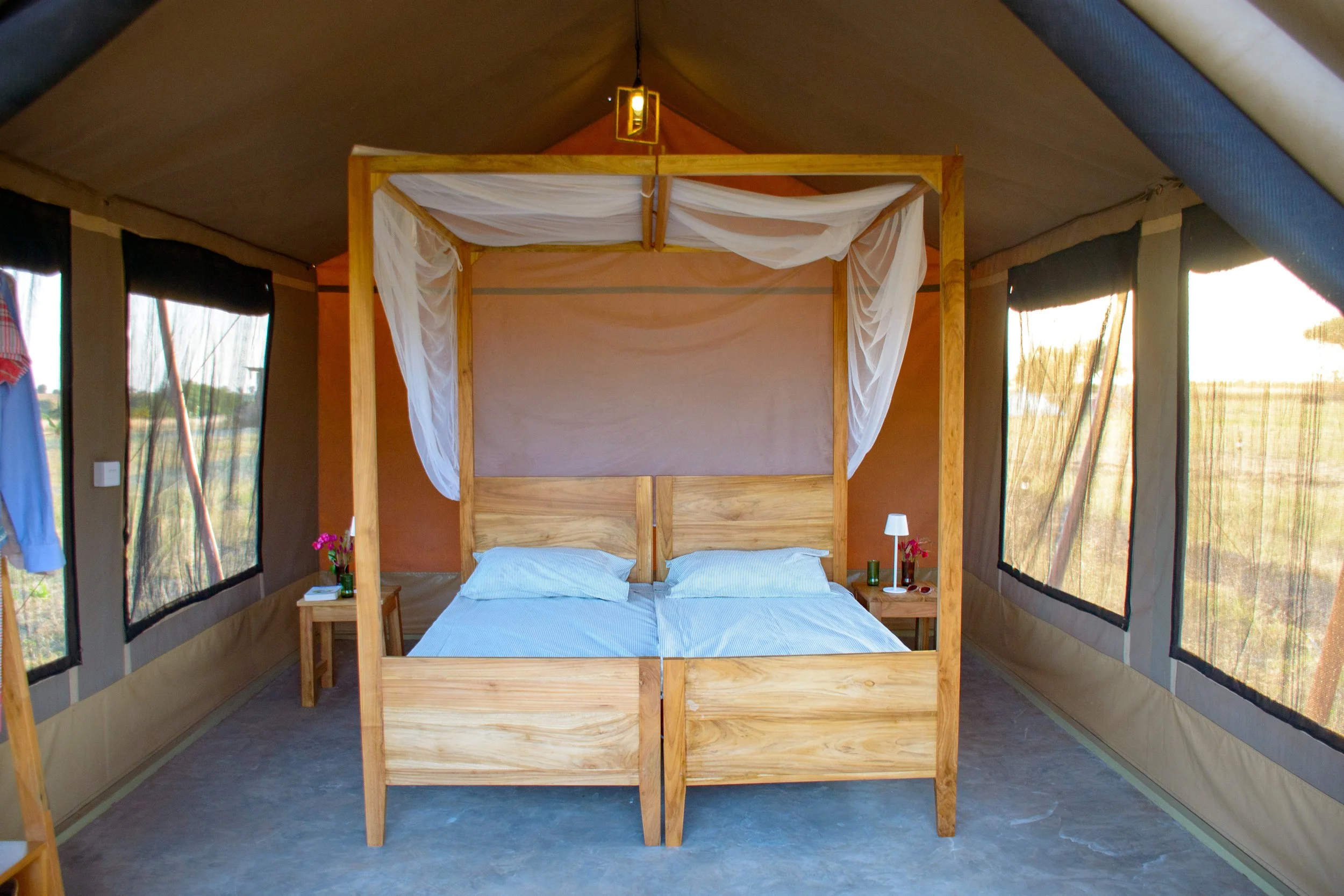 Interior of a tent with a wooden bed frame and white canopy drapes, two beds with white sheets, dark curtains on the sides, and small nightstands with lamps at Njiani Camp, Moshi.