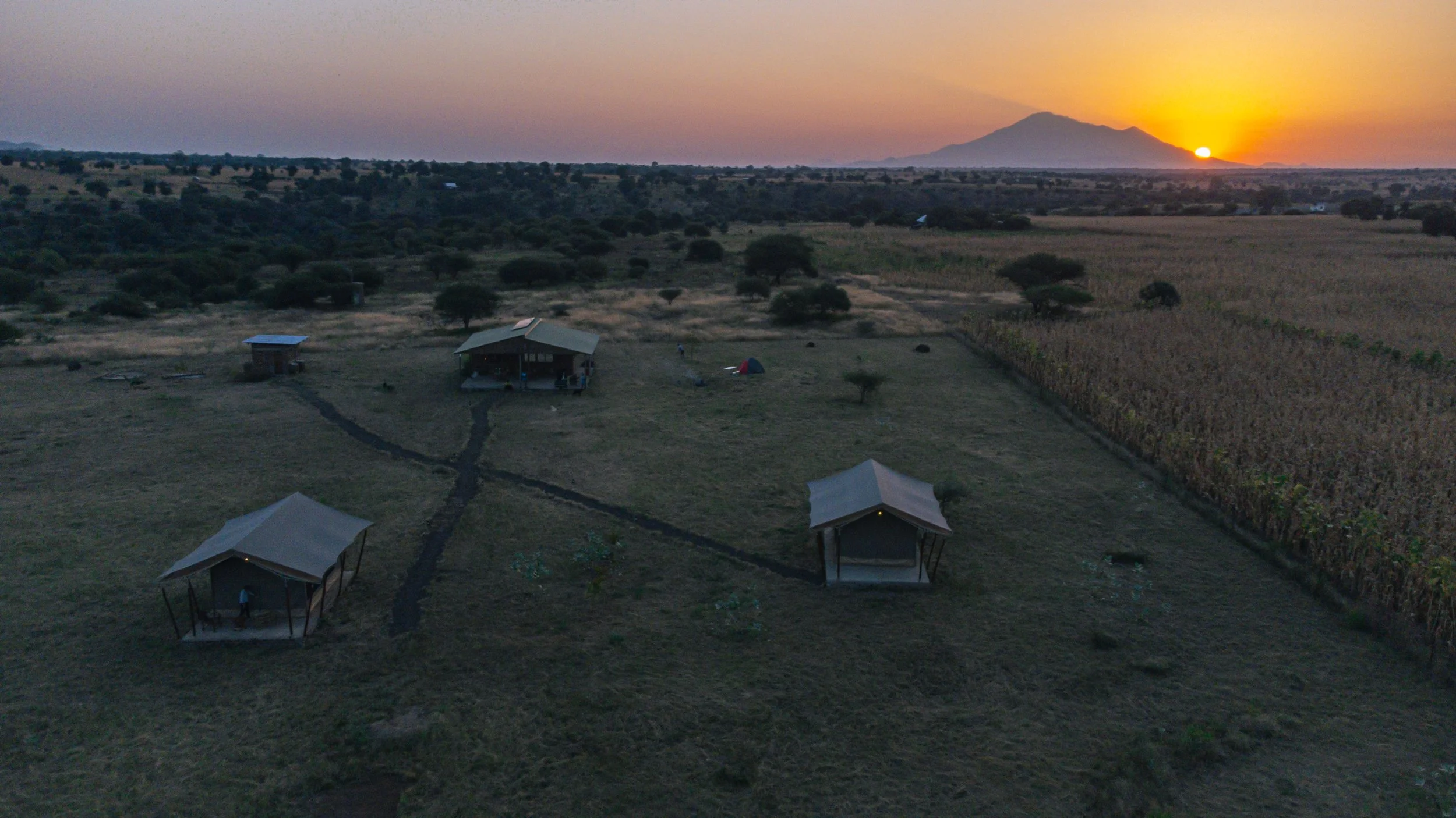 An aerial view of Njiani River Camp at sunset with small huts, tents, and walking paths in an open field. In the background, there are trees, farmland, and a mountain with the sun setting behind it.
