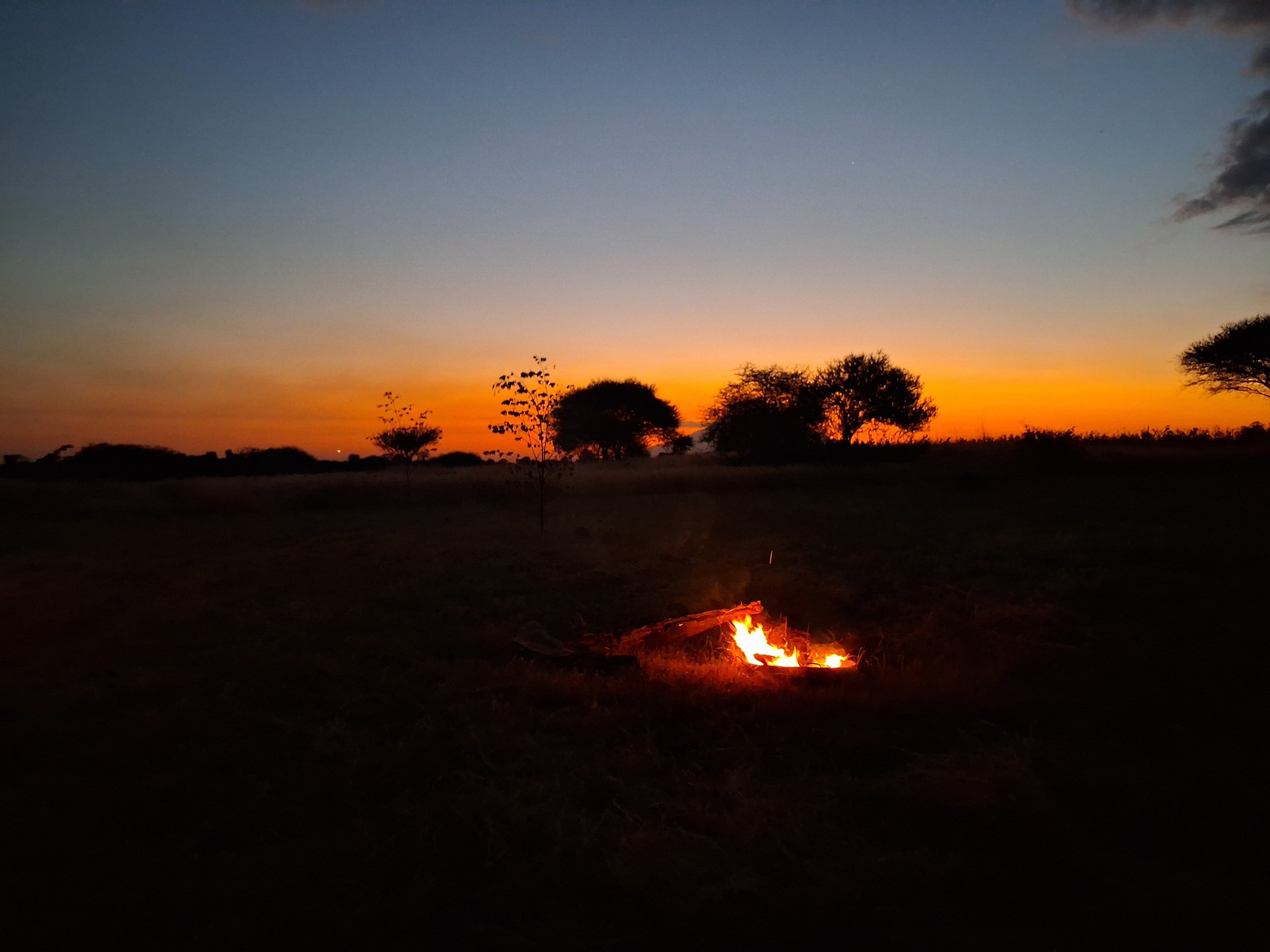 Sunset or sunrise over Njiani River Camp There is a small fire burning on the ground in the foreground.