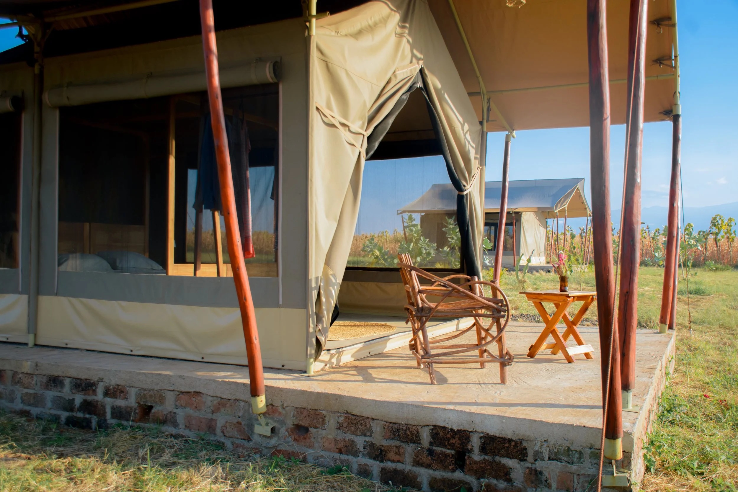View of a glamping tent at Njiani River Camp, outside of Moshi, on a raised brick platform, with outdoor furniture including a wooden chair and a small table, set in a rural area with fields and a distant building under a clear sky.