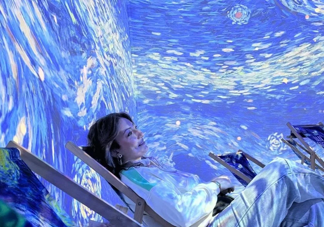 Une femme confortablement installée dans une chaise pliable, entourée d'une projection immersive de l'œuvre 'Nuit étoilée' de Vincent van Gogh sur les murs et le plafond, créant une atmosphère artistique et relaxante.