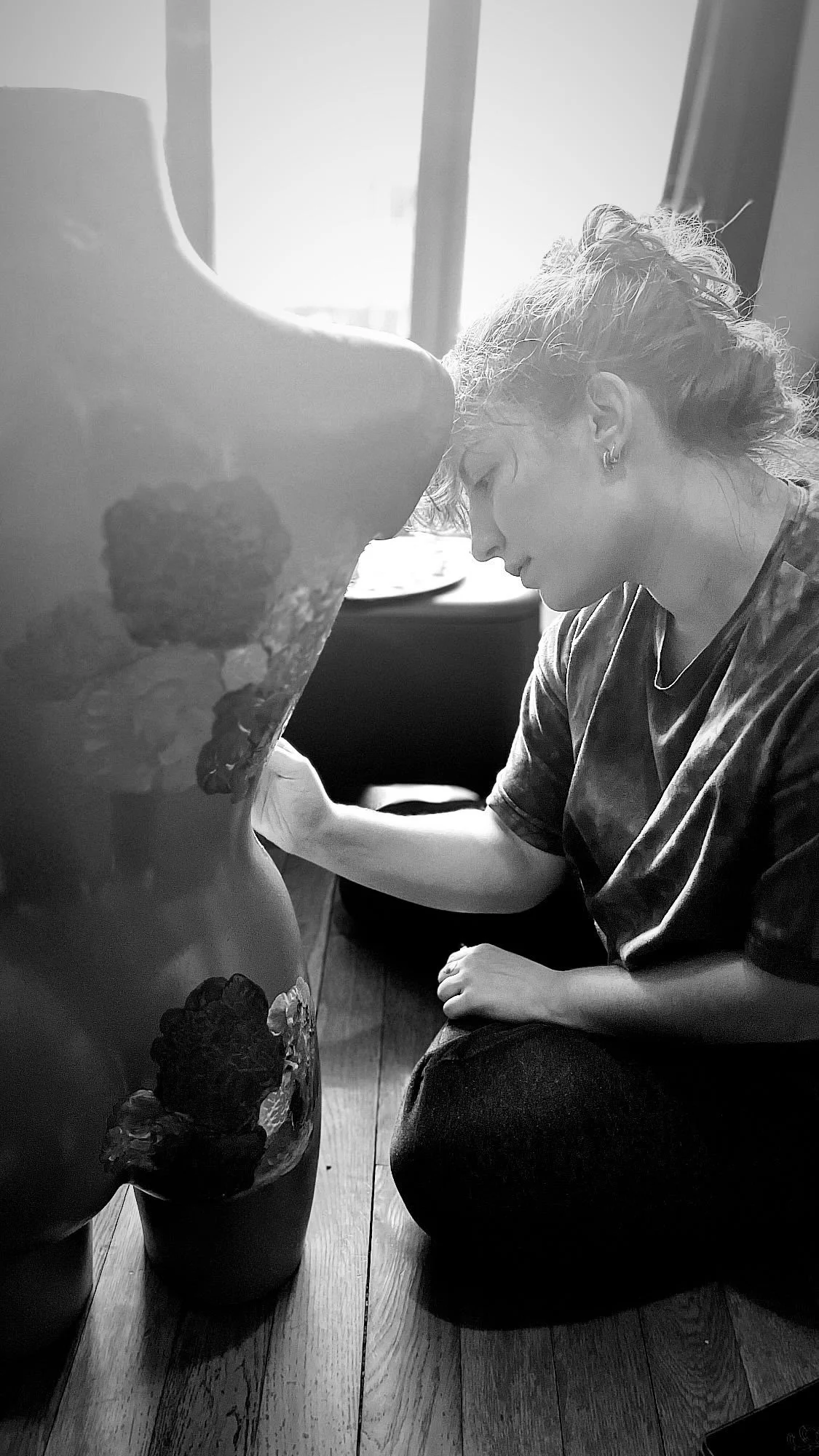 Une femme est en train de peindre un mur ou une surface avec des fleurs, en position accroupie. La photo est en noir et blanc.