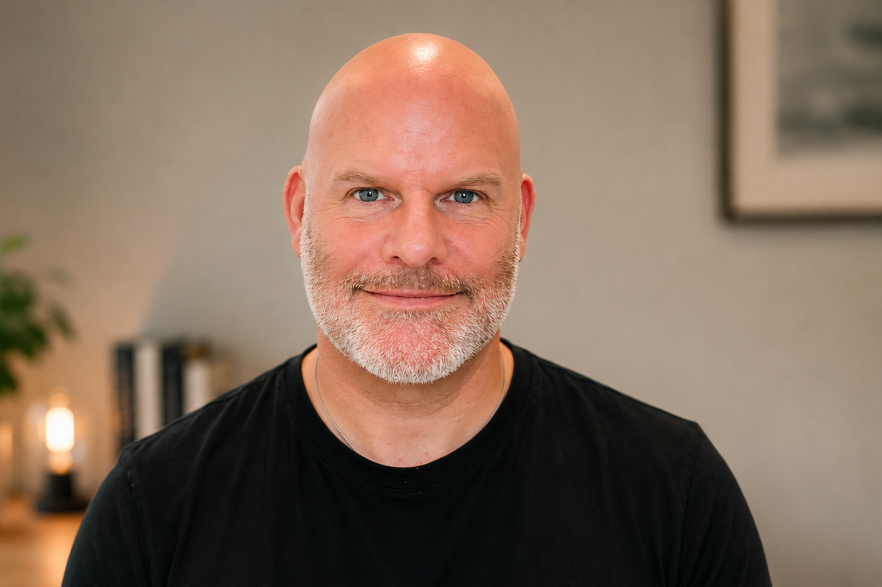 A middle-aged man with a bald head, blue eyes, and a beard, wearing a black shirt, smiling at the camera in an indoor setting.