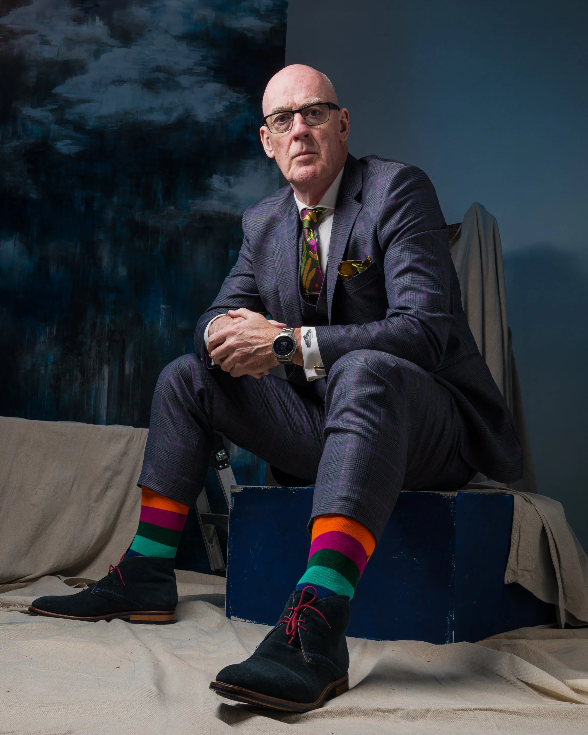A bald man wearing glasses, a checkered suit, colorful striped socks, and black shoes sitting on a chair in an art studio or gallery. He has a serious expression and is looking at the camera. Behind him are dark blue and black abstract paintings, and the floor is covered with a beige fabric.