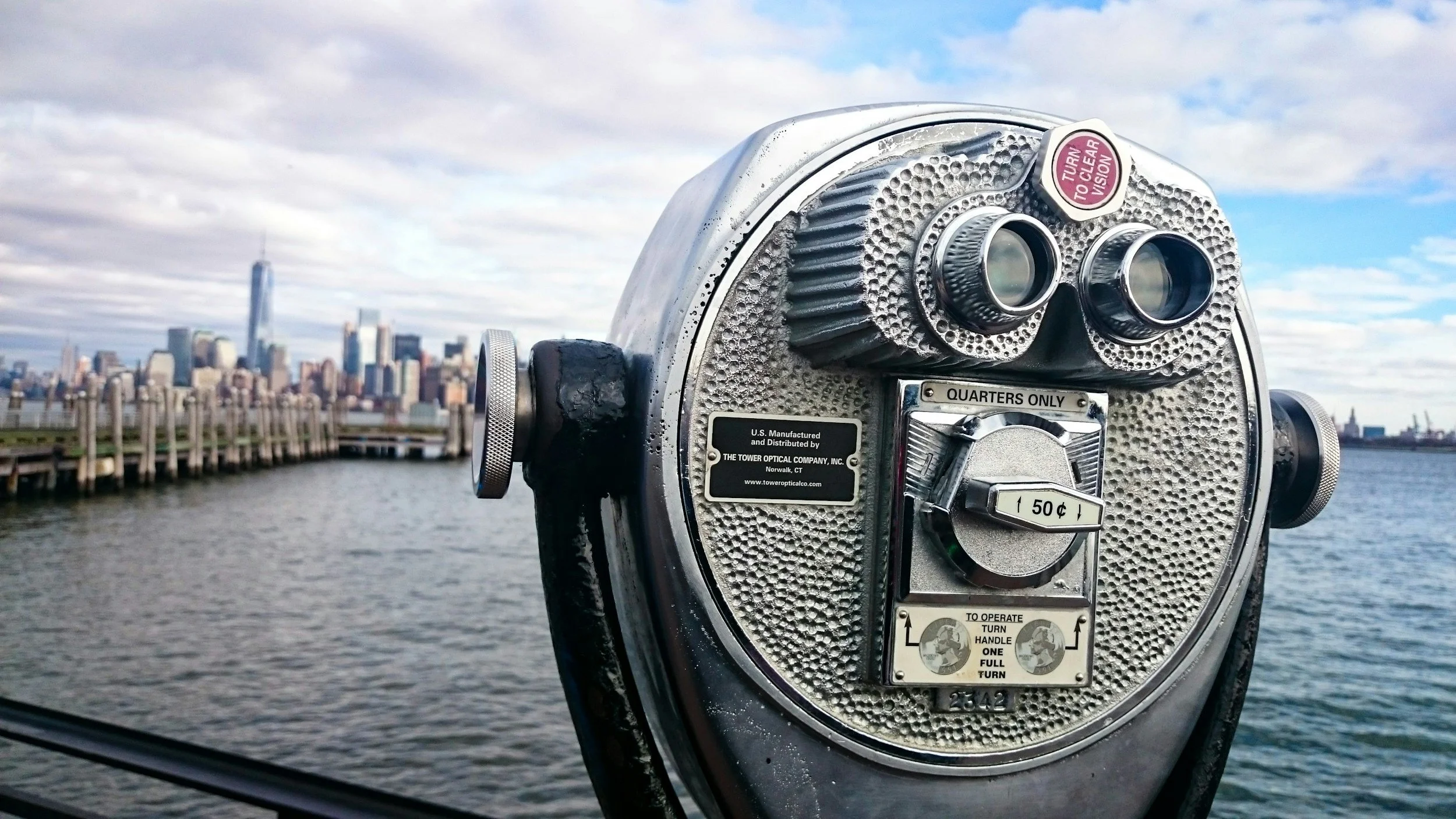 A coin-operated viewing binoculars station with a city skyline and water in the background, cloudy sky.