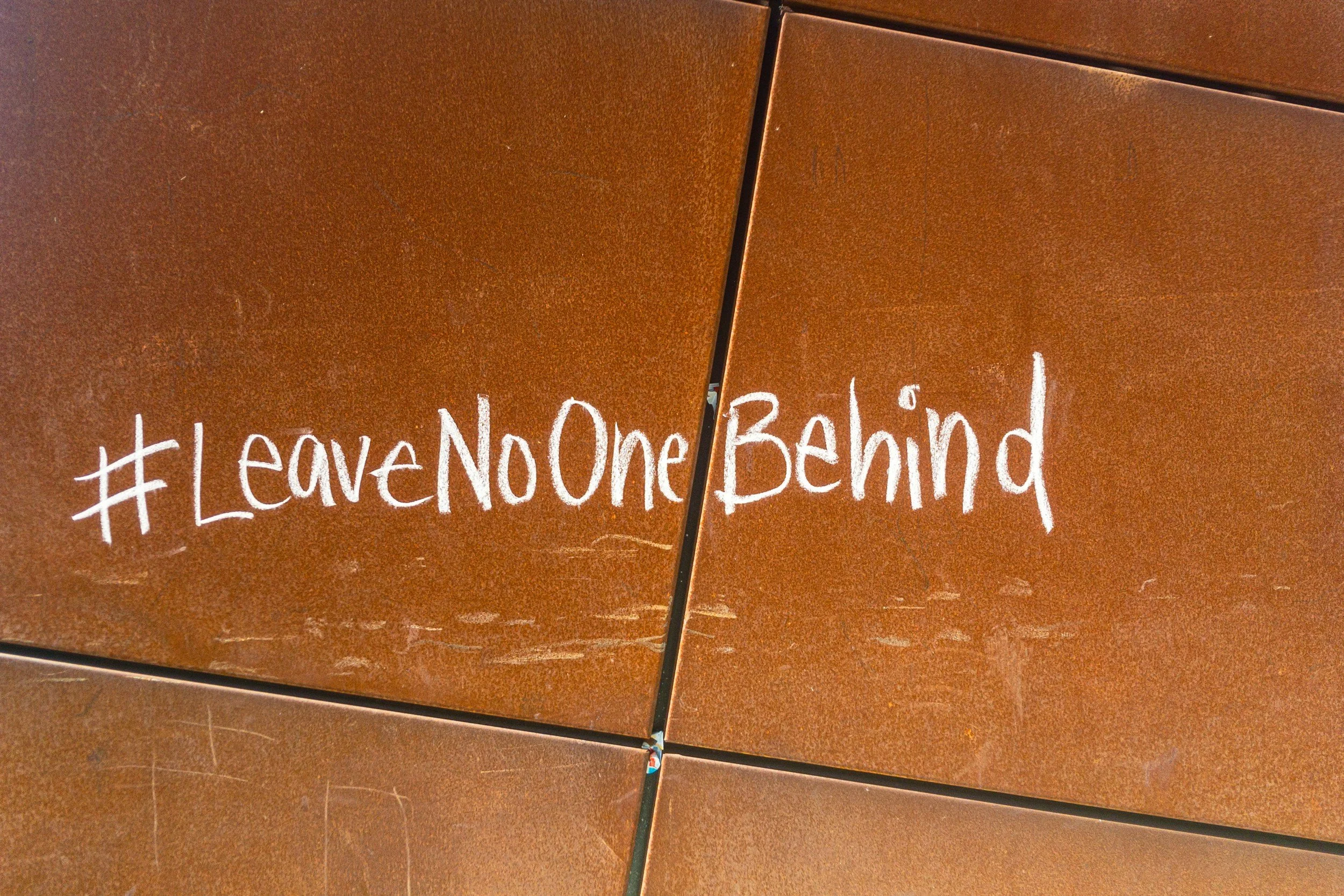 Close-up view of a brown, textured surface with a handwritten message in white chalk that reads #LeaveNoOneBehind.