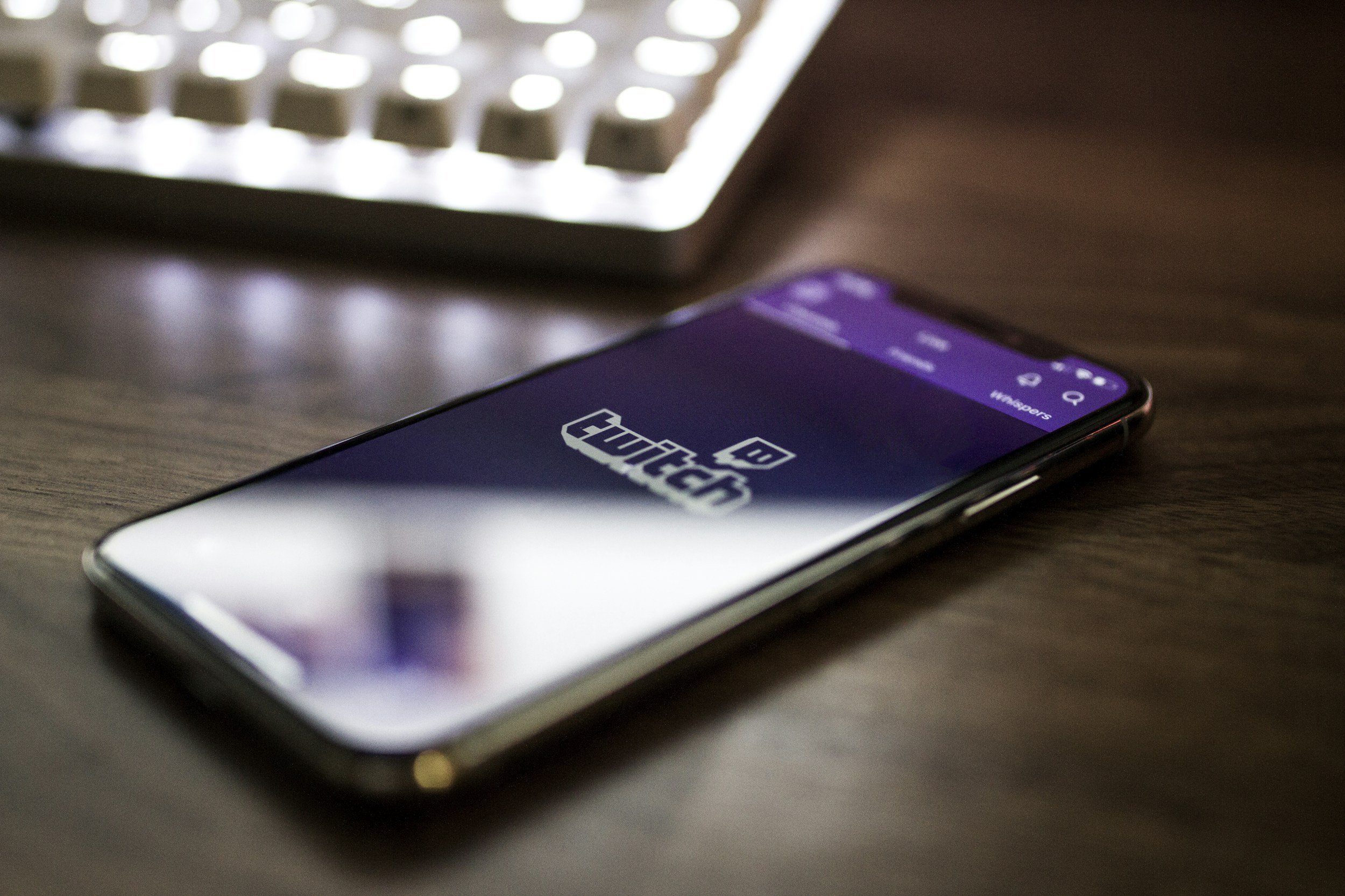 Close-up of a smartphone on a table with a Twitch app open on the screen, showing the Twitch logo and interface, with a blurred keyboard in the background.