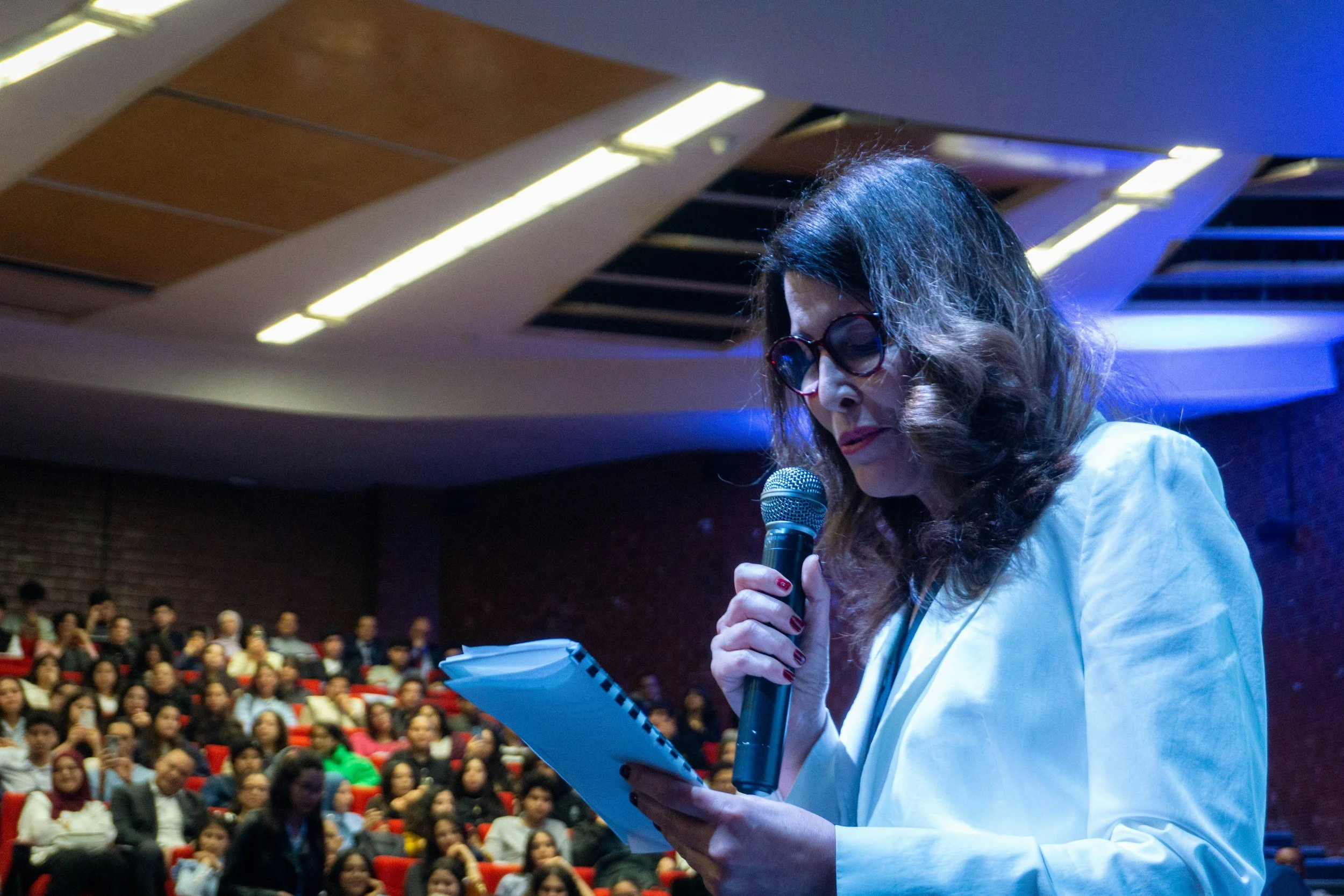 A woman with wavy brown hair, wearing glasses and a white blazer, is speaking into a microphone while looking at notes or a script. She is at a podium in front of an audience in a large auditorium with a sloped ceiling and bright lights.