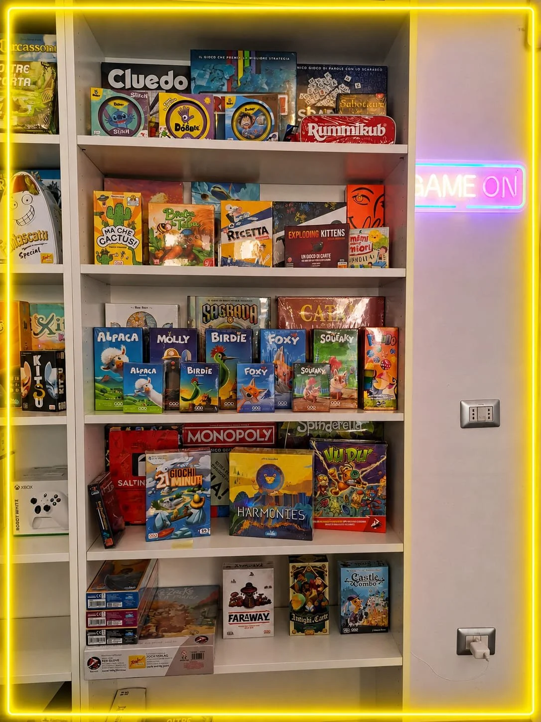 Mensola con vari giochi da tavolo e libri illustrati per bambini, tra cui Monopoly, Sacrago, Vudu, e giochi di carte, con neon rosa e blu sul muro